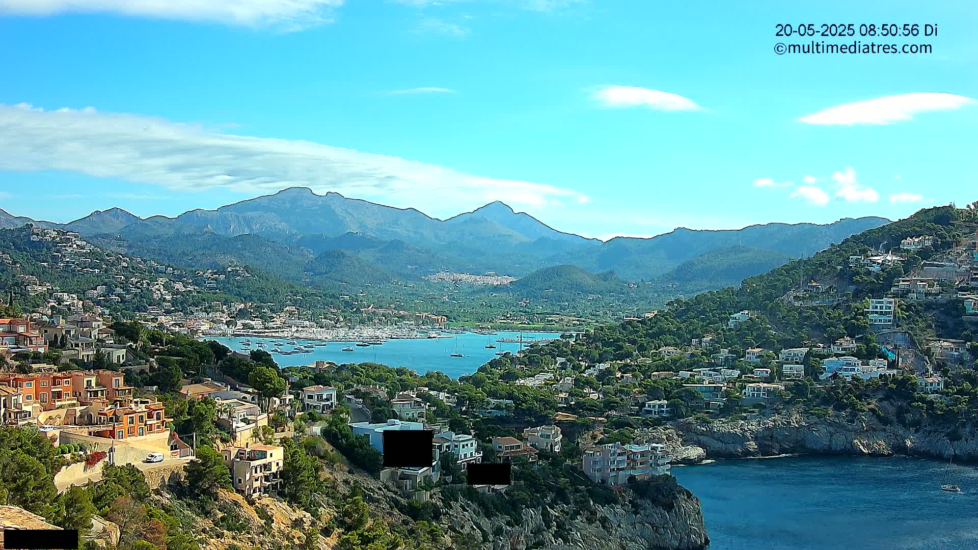 A coastal town nestled against a mountain range under a mostly sunny sky with a few scattered clouds, featuring a harbor filled with sailboats.