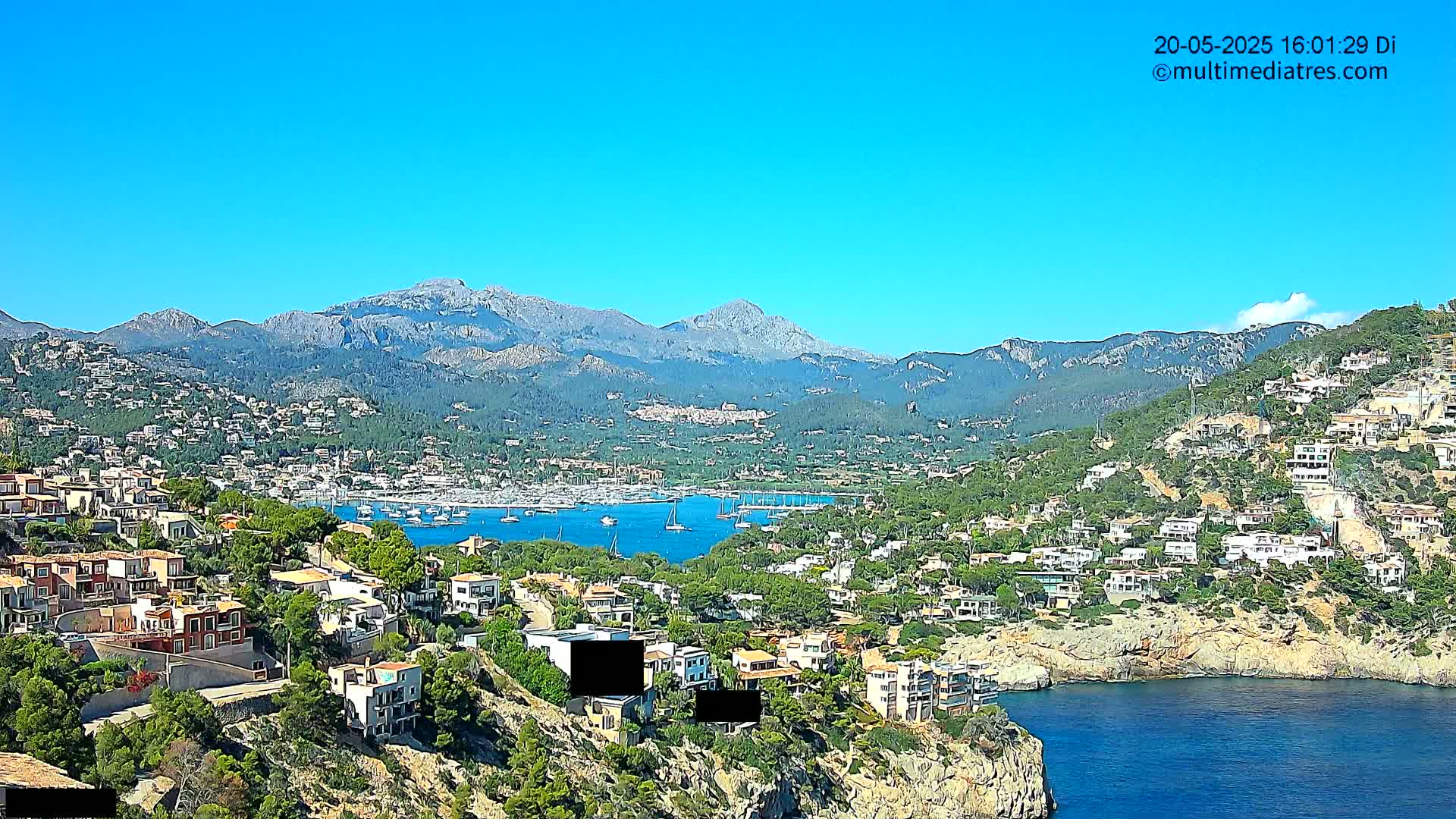 A coastal town nestled in a mountainous region with a clear blue sky and a calm harbor filled with sailboats.