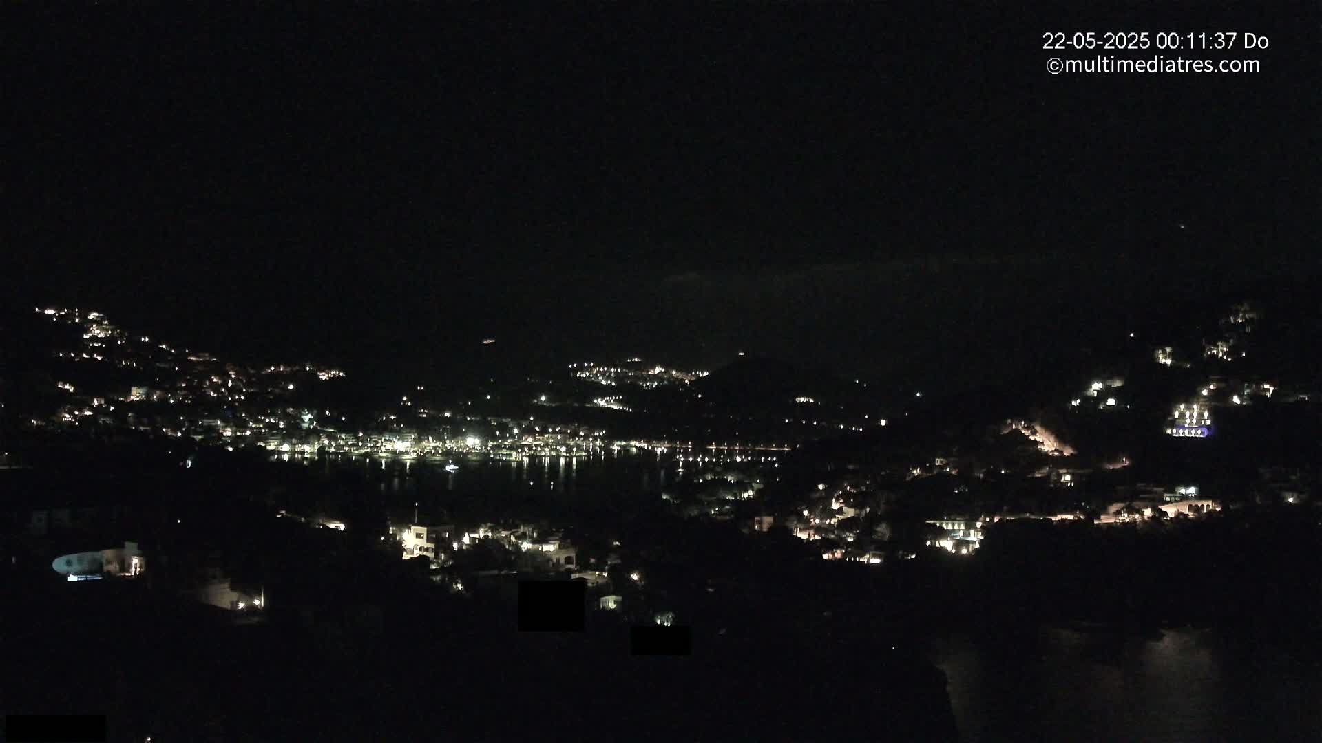 A nighttime view of a coastal town nestled on hillsides, with scattered lights illuminating the buildings and a dark body of water reflecting some of the lights.