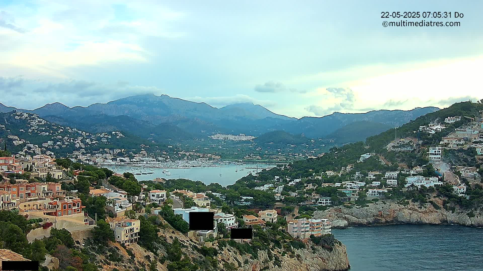 A coastal town nestled in a mountainous region, with a calm bay filled with boats under a partly cloudy sky.