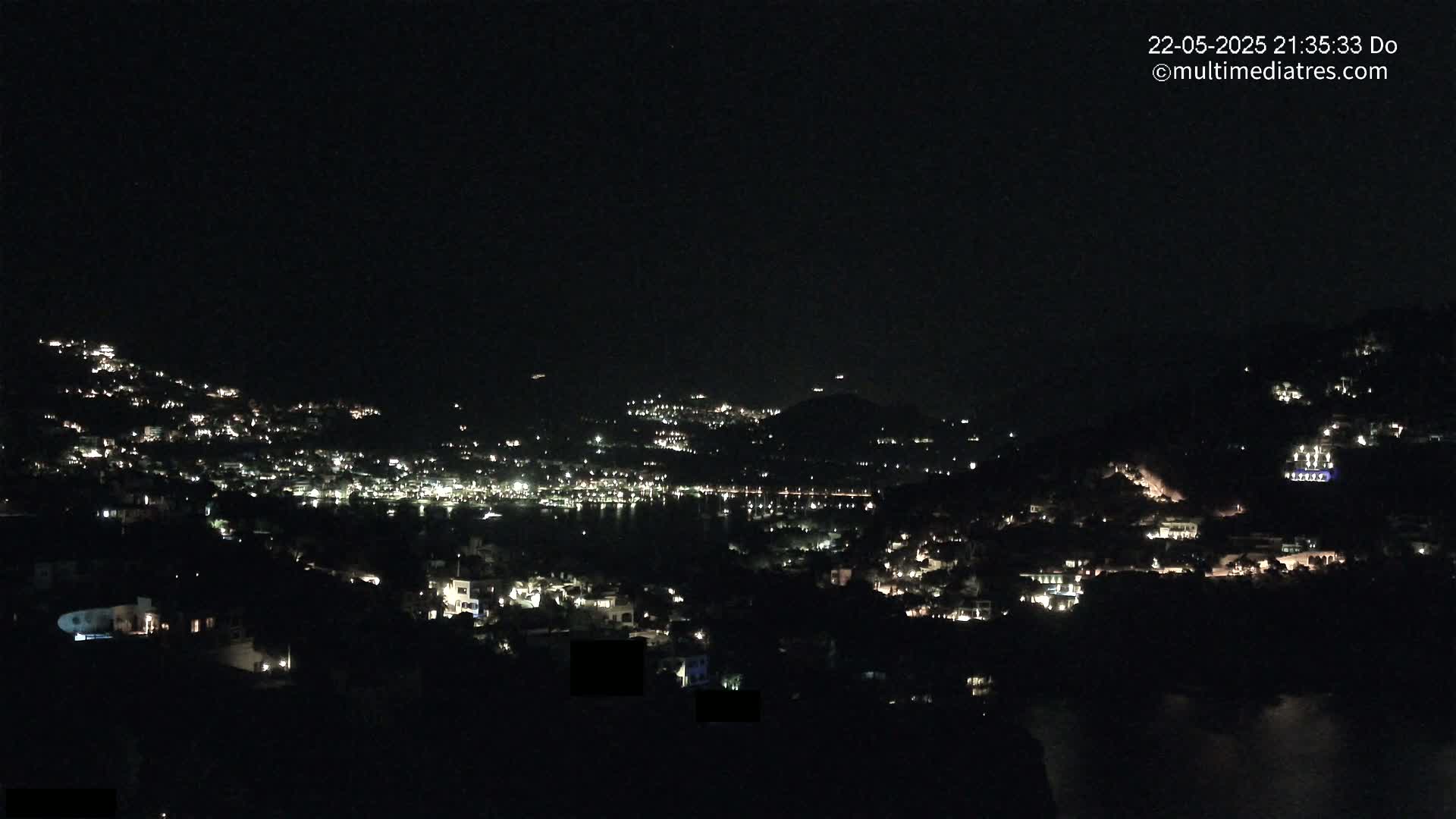 A nighttime view of a coastal town nestled on hillsides, with numerous lights illuminating the buildings and surrounding water.