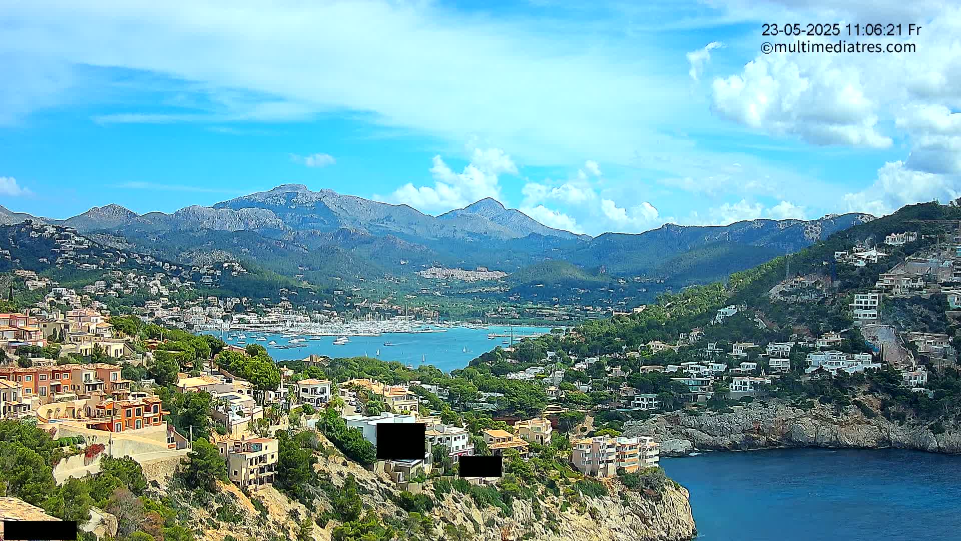 A coastal town nestled in a mountainous region, with a harbor filled with boats under a partly cloudy sky.
