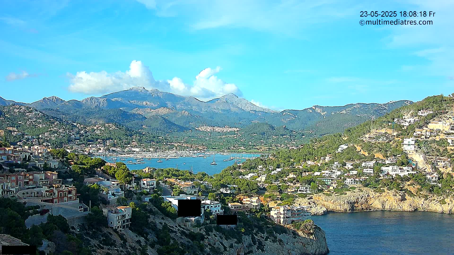 A coastal town nestled in a mountainous region under a mostly sunny sky with some scattered clouds, features a harbor filled with boats.