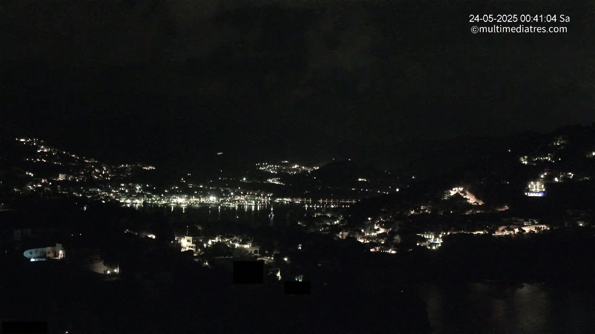A nighttime view of a coastal town nestled on hillsides, illuminated by scattered lights, under a dark, cloudy sky.