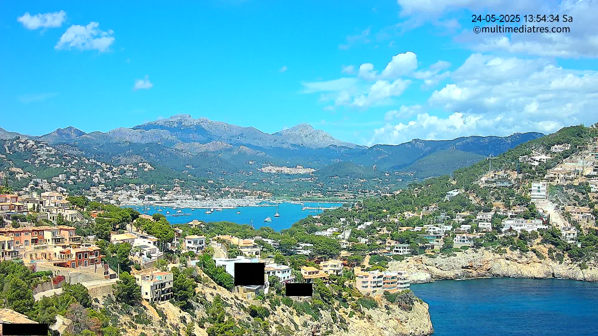 A coastal town nestled between mountains under a mostly sunny sky with scattered clouds overlooks a calm bay filled with sailboats.
