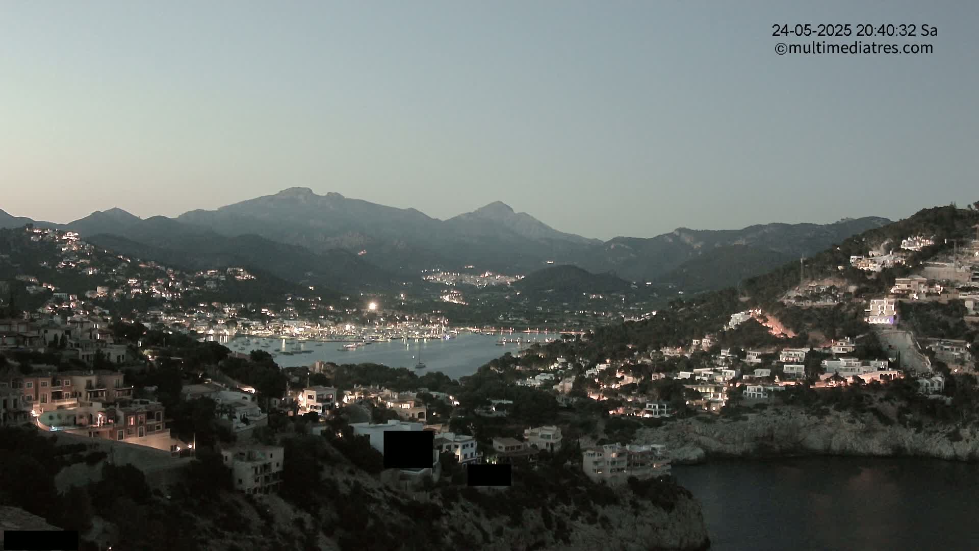 A nighttime view of a coastal town nestled among hills, illuminated by lights, with a calm body of water in the foreground and mountains in the background under a twilight sky.