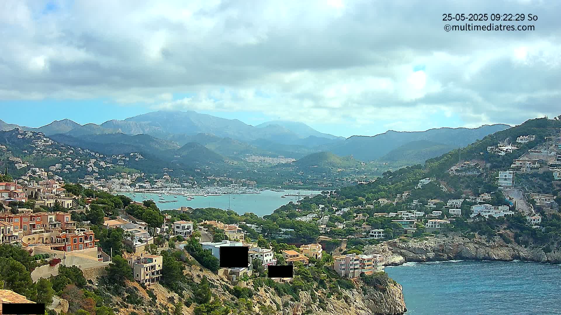 A coastal town nestled in mountains overlooking a calm bay filled with boats under a partly cloudy sky.