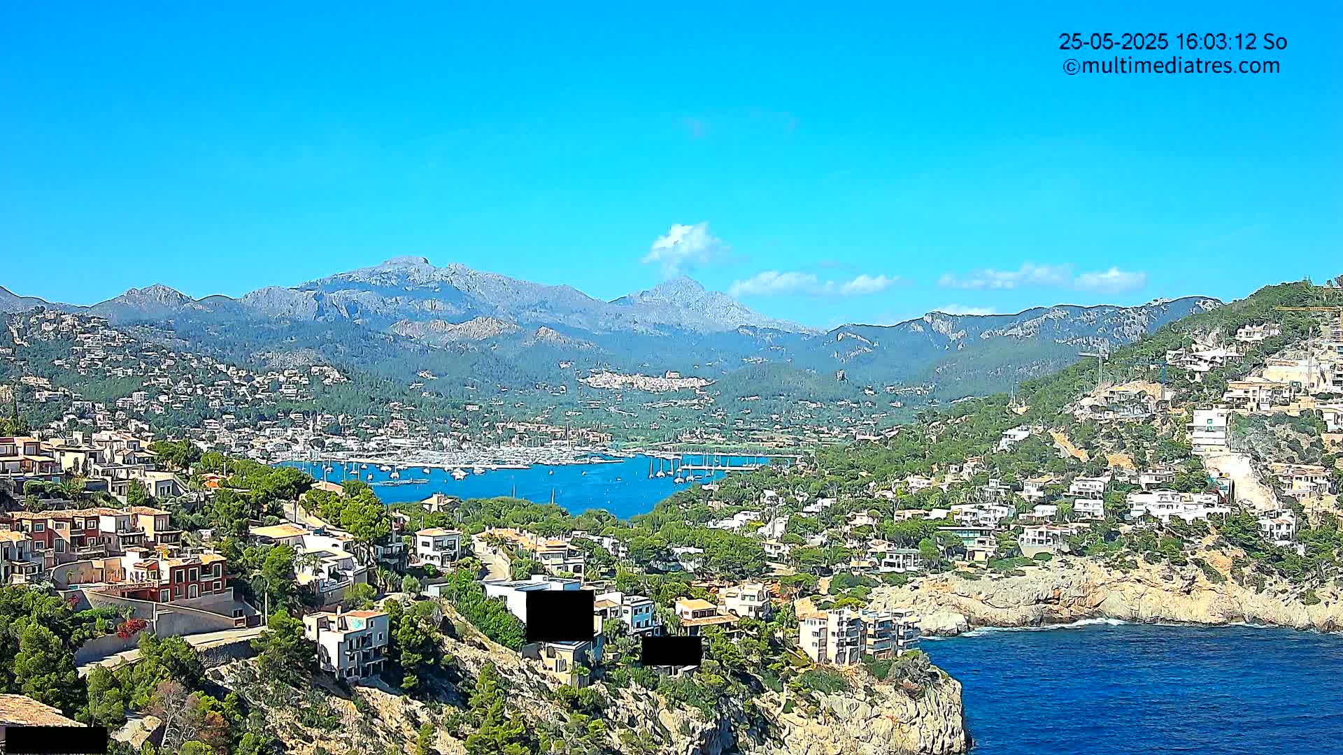 A coastal town nestled on a hillside overlooking a harbor filled with boats, with mountains in the background under a clear blue sky.