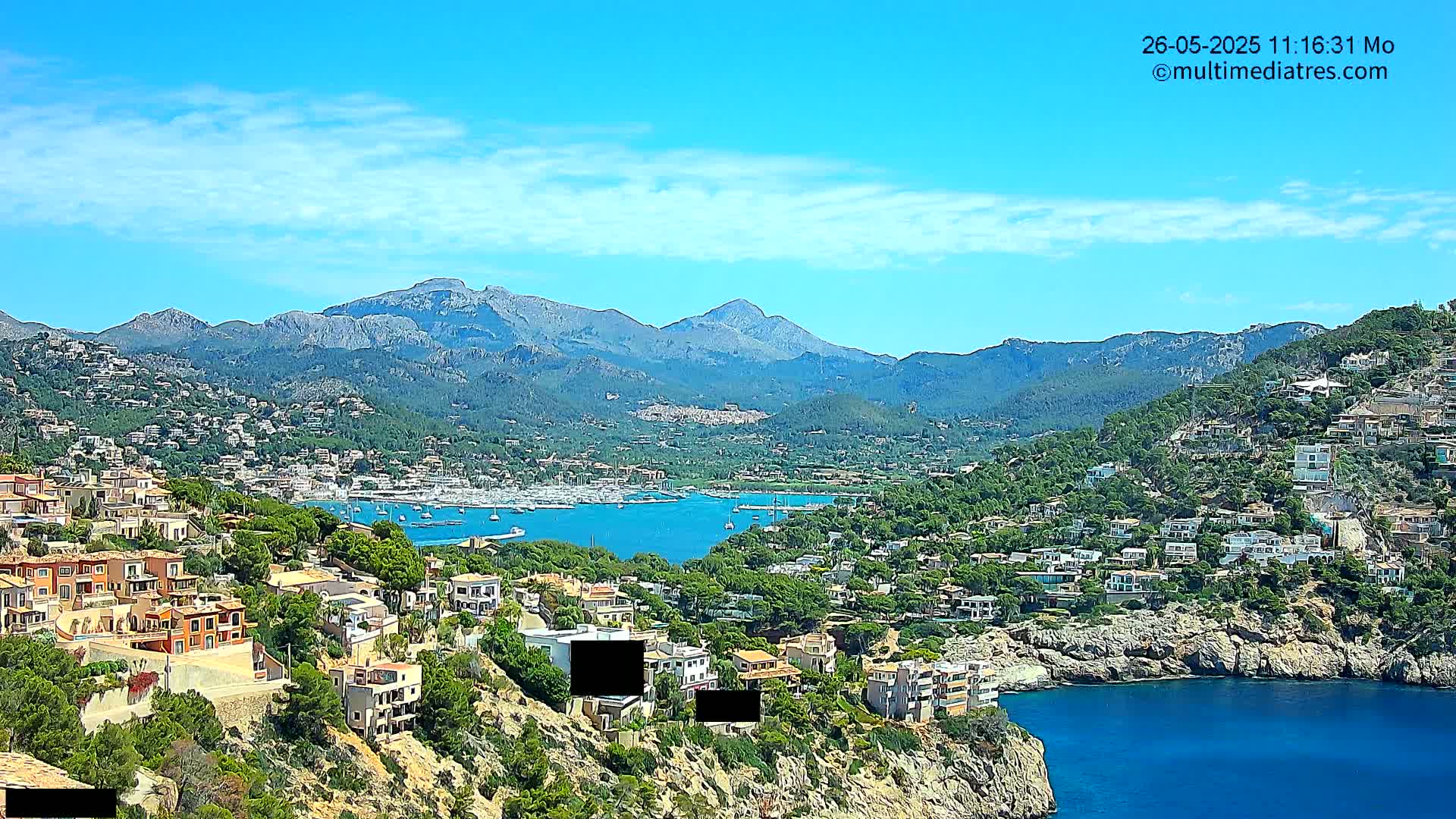 A coastal town nestled in a mountainous region, under a clear blue sky with scattered clouds, features a calm bay filled with boats.