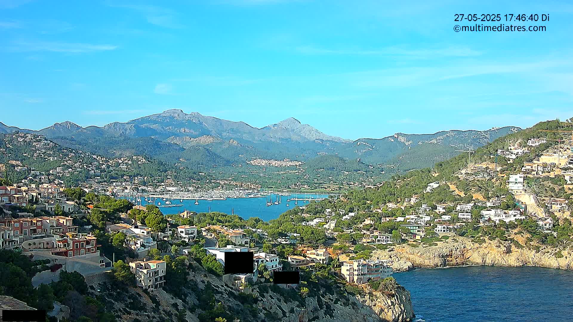 A coastal town nestled against a mountain range under a clear blue sky, with a harbor full of sailboats visible in the distance.