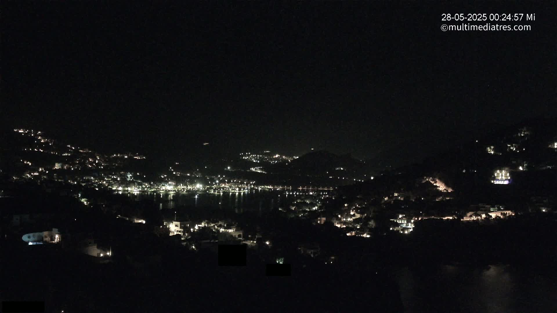 A nighttime view shows a coastal town nestled on hillsides, with many lights illuminating the buildings and a dark body of water separating them.