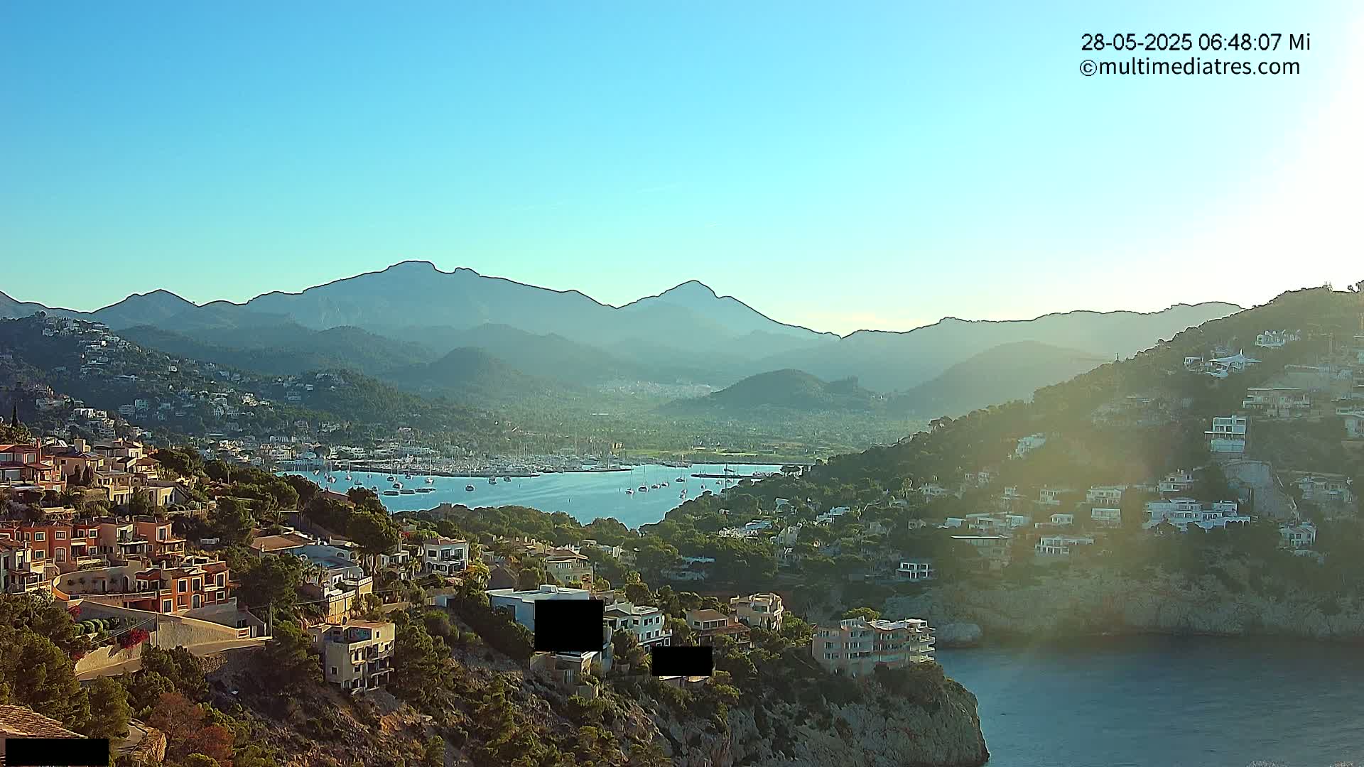 A coastal town nestled on a hillside overlooking a calm bay with numerous boats, all set against a backdrop of sunlit mountains under a clear blue sky.