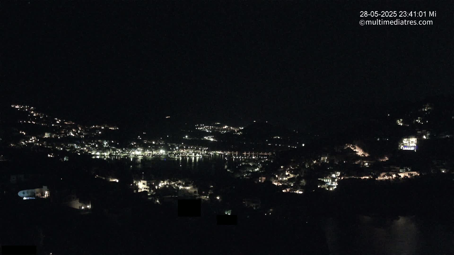 A nighttime, aerial view shows a coastal town with scattered lights along hillsides and a dark body of water.