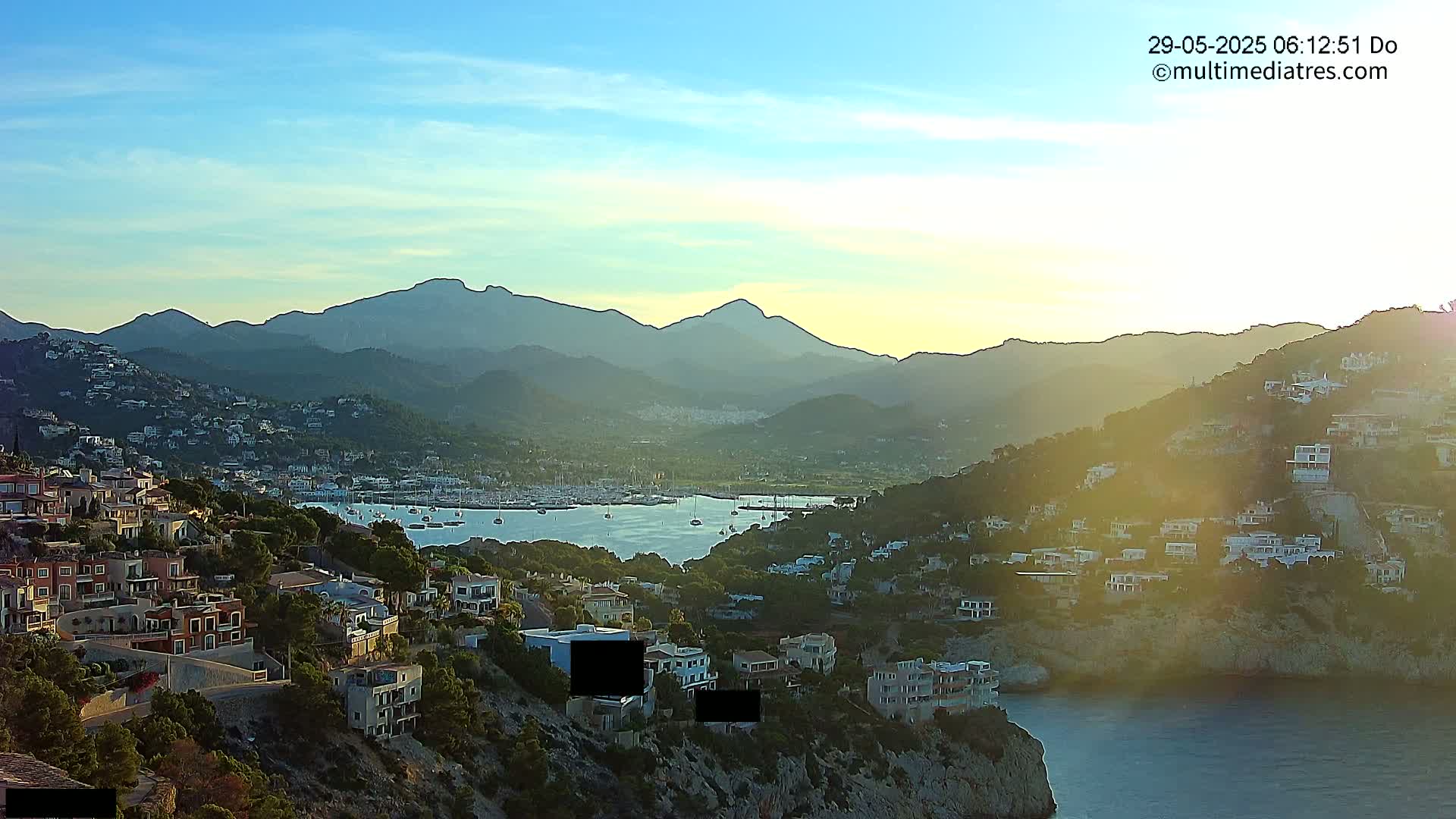 A coastal town nestled against a mountain range, with a harbor filled with boats, under a clear, sunset sky.
