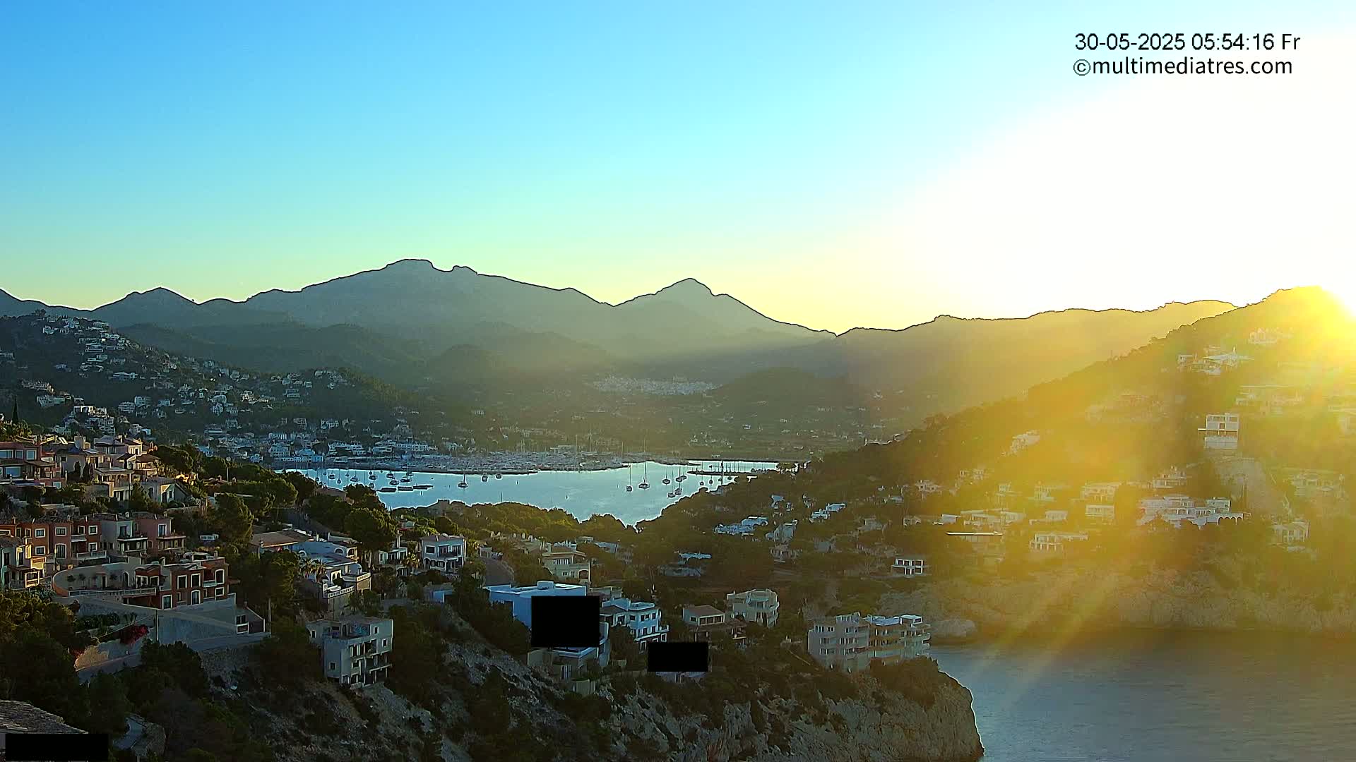 A coastal town nestled in a hillside overlooking a calm bay filled with boats, all bathed in the golden light of a sunrise or sunset, with mountains in the background.