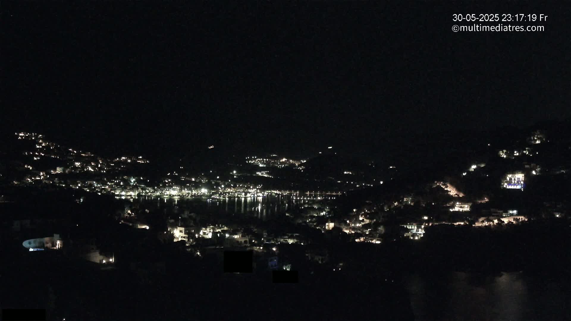 A nighttime, long-range view of a town nestled on a hillside, with its lights illuminating the dark landscape and reflecting on the water below.