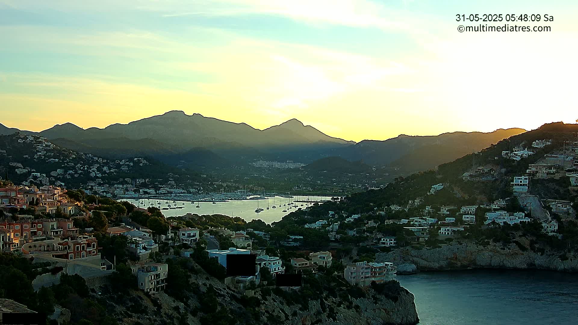 A coastal town nestled among hills at sunset, with a calm bay filled with sailboats in the foreground and mountains in the background.