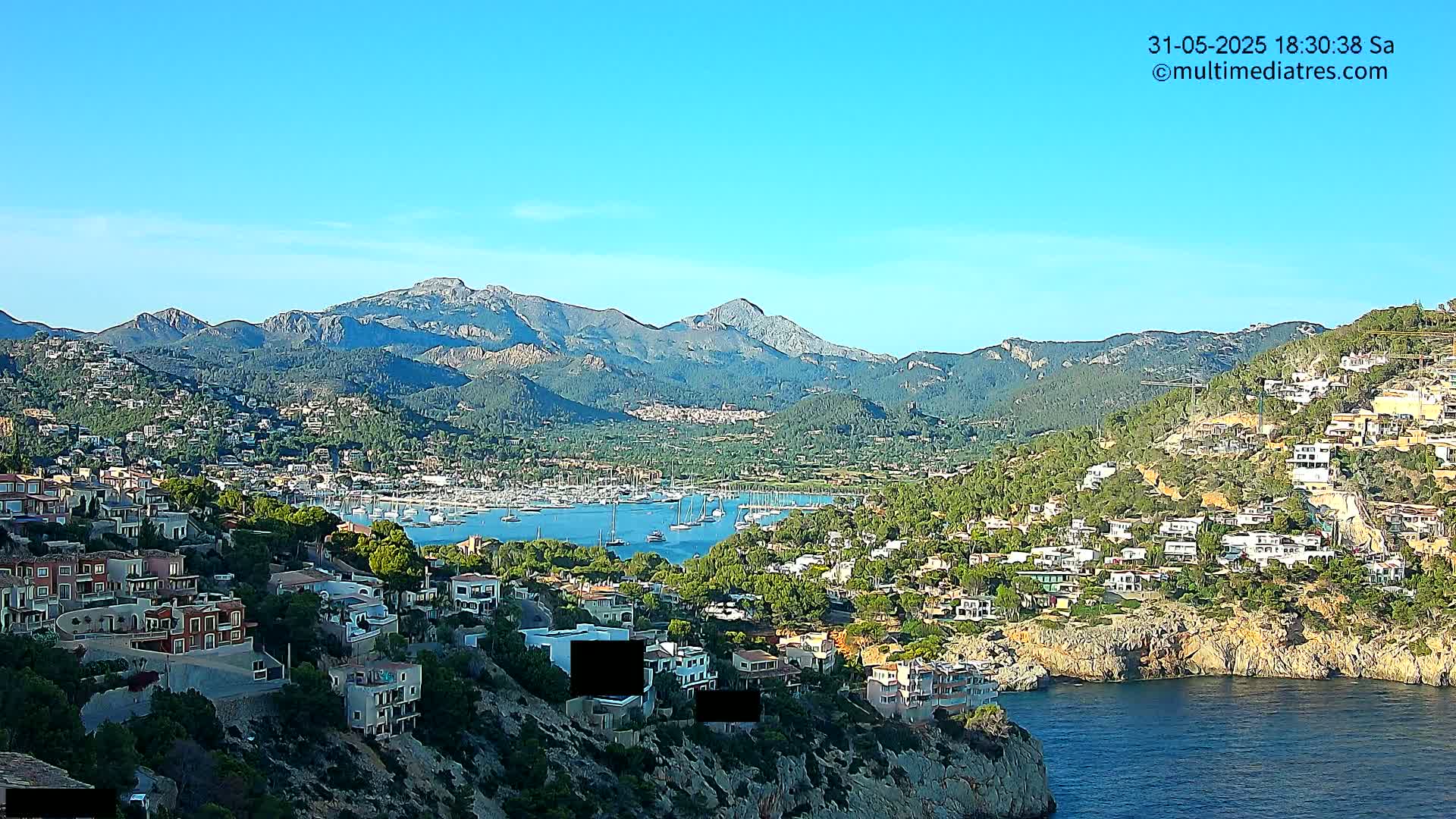 A coastal town nestled below mountains, featuring a harbor filled with sailboats, under a clear blue sky.