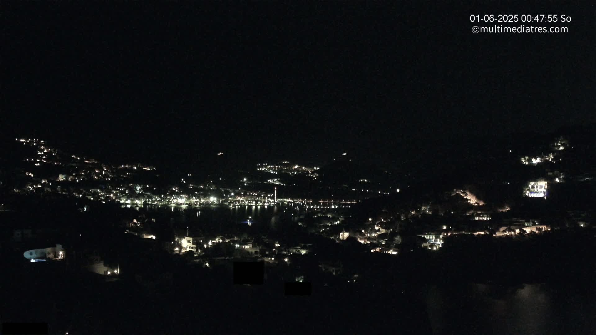 A nighttime, long-range view of a coastal town illuminated by scattered lights, with a dark body of water visible in the middle ground.