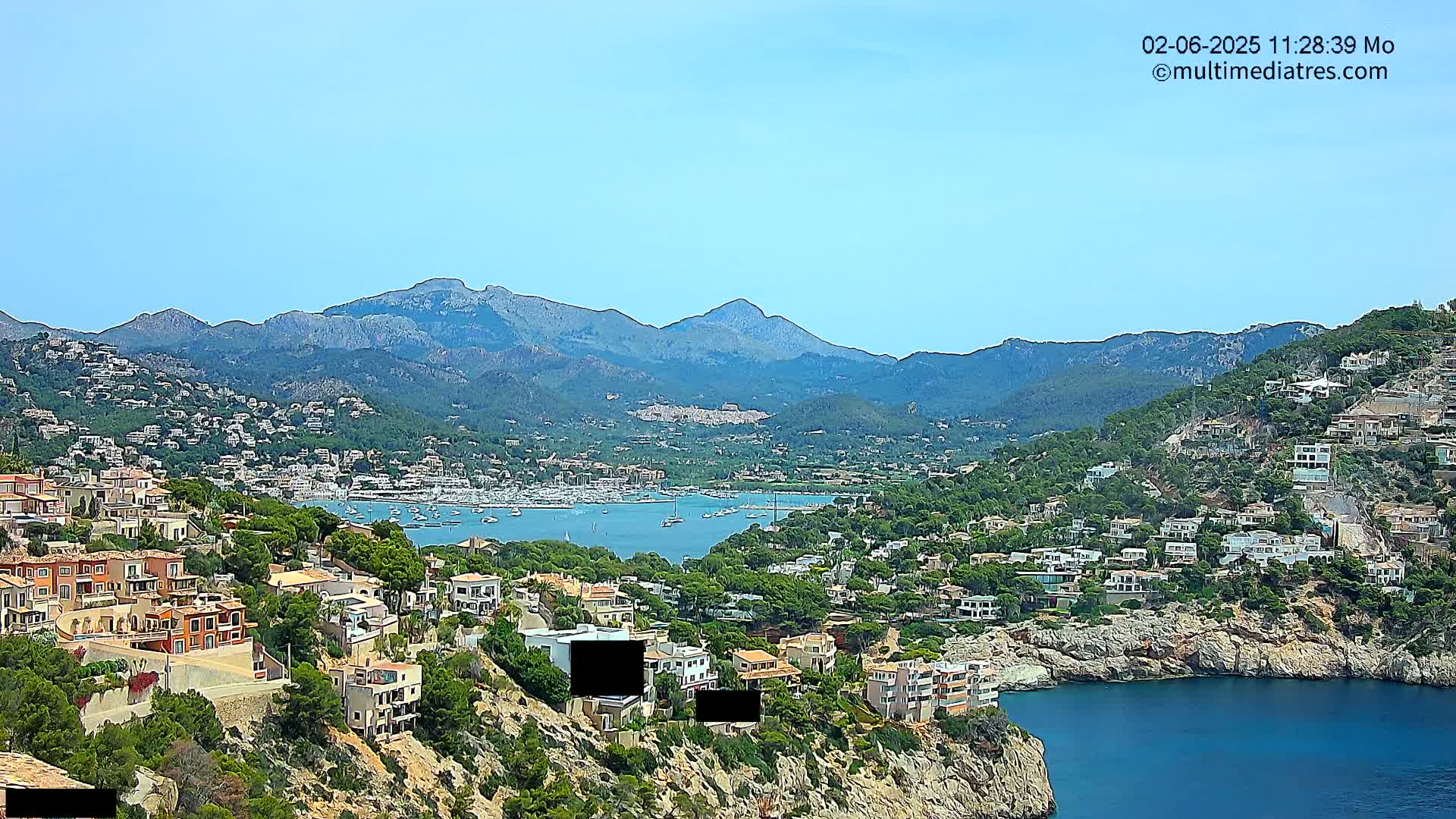 A coastal town nestled among hills and mountains on a clear, sunny day, with a calm bay filled with boats visible in the distance.