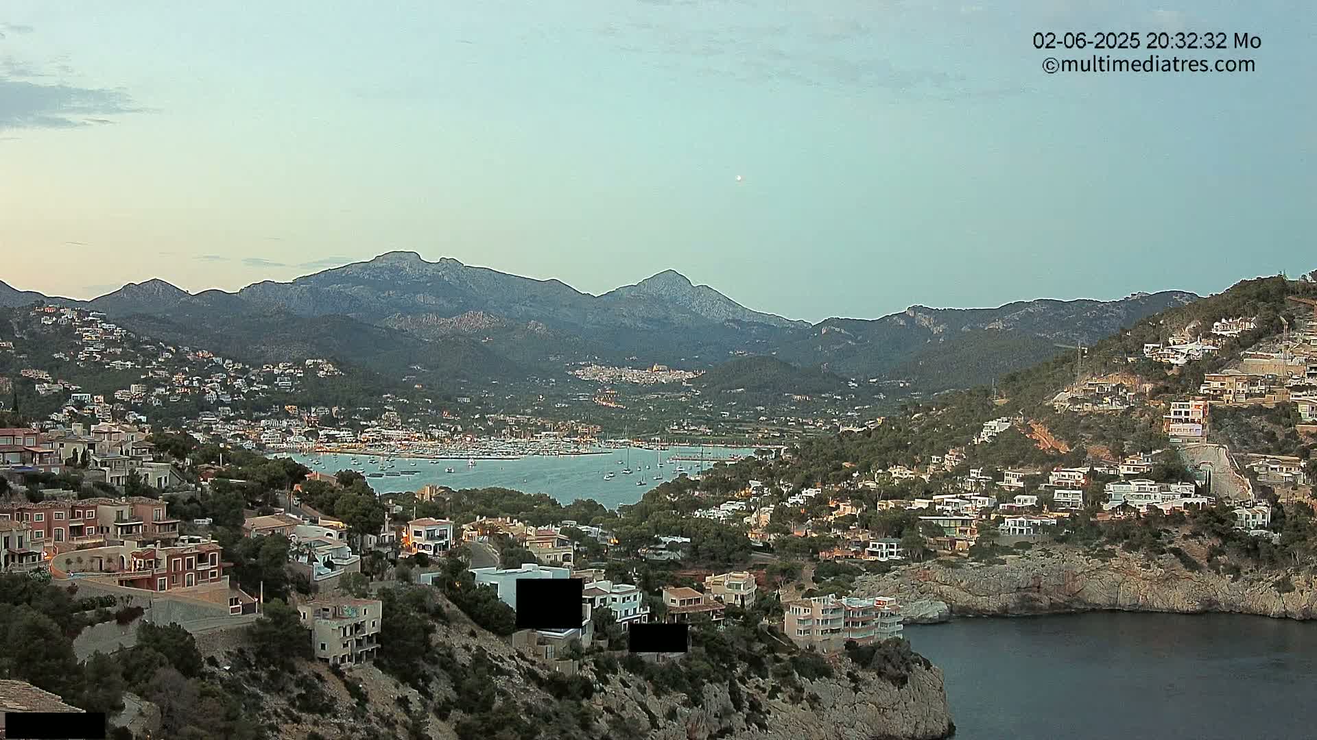 A coastal town nestled between hills and mountains, overlooking a calm bay filled with boats under a clear twilight sky.