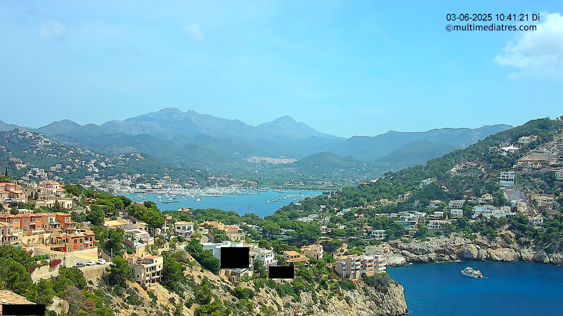 A coastal town nestled in a mountainous region on a clear, sunny day, with a harbor filled with boats visible in the distance.