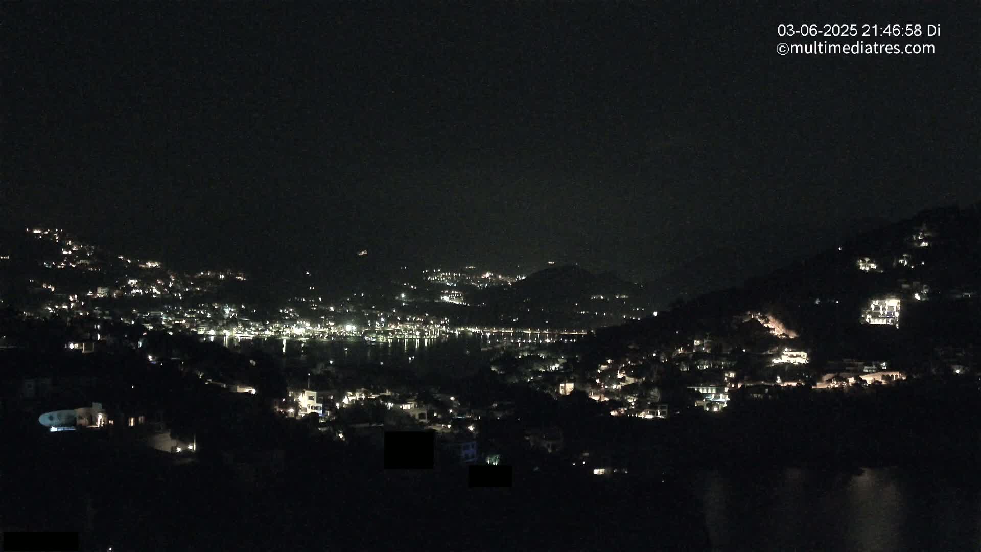 A nighttime view shows a coastal town with scattered lights illuminated across hillsides and along a dark body of water under a clear, dark sky.