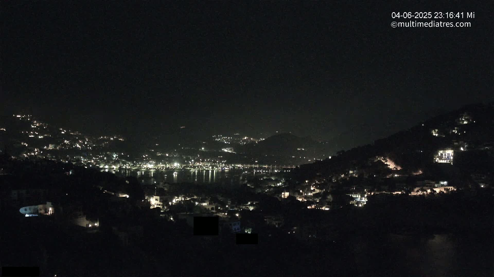 A nighttime view of a coastal town nestled on hillsides, with scattered lights illuminating the buildings and a dark, calm body of water reflecting the lights.
