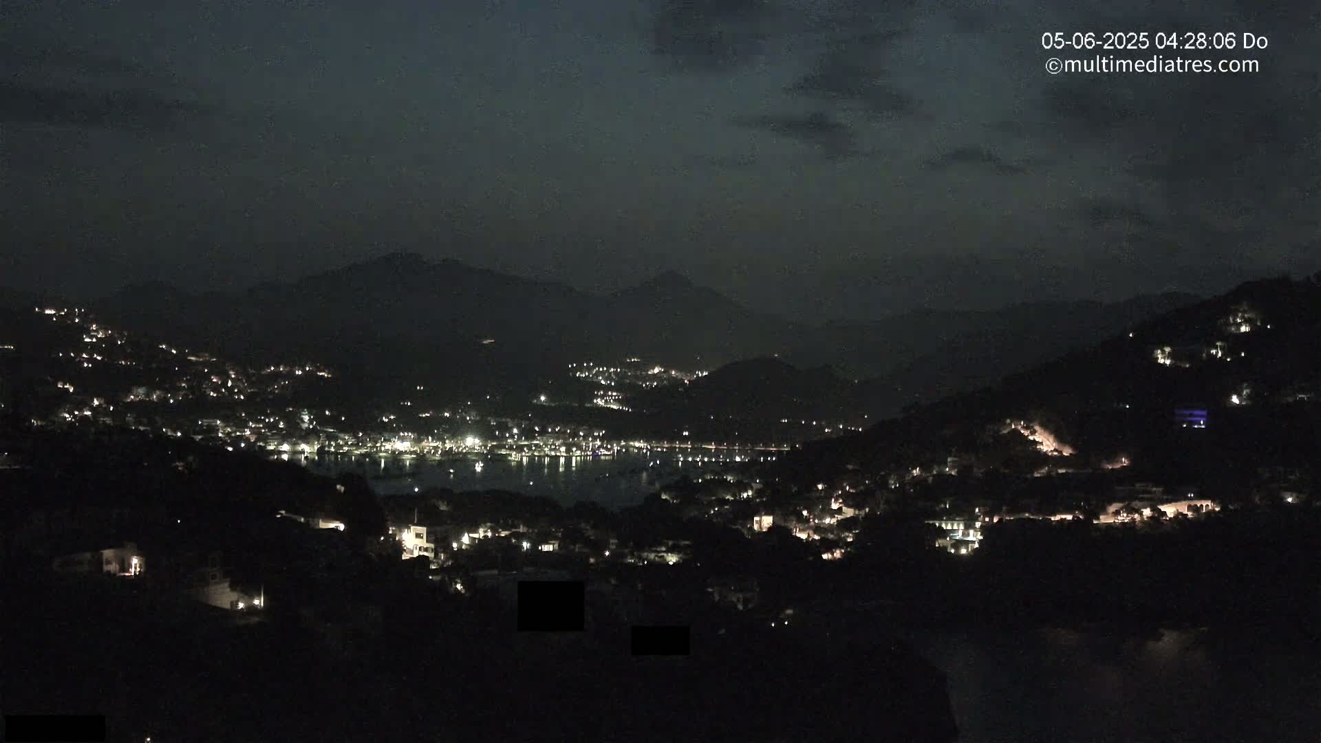 A nighttime view of a coastal town nestled in dark hills, illuminated by scattered lights, under a partly cloudy sky.