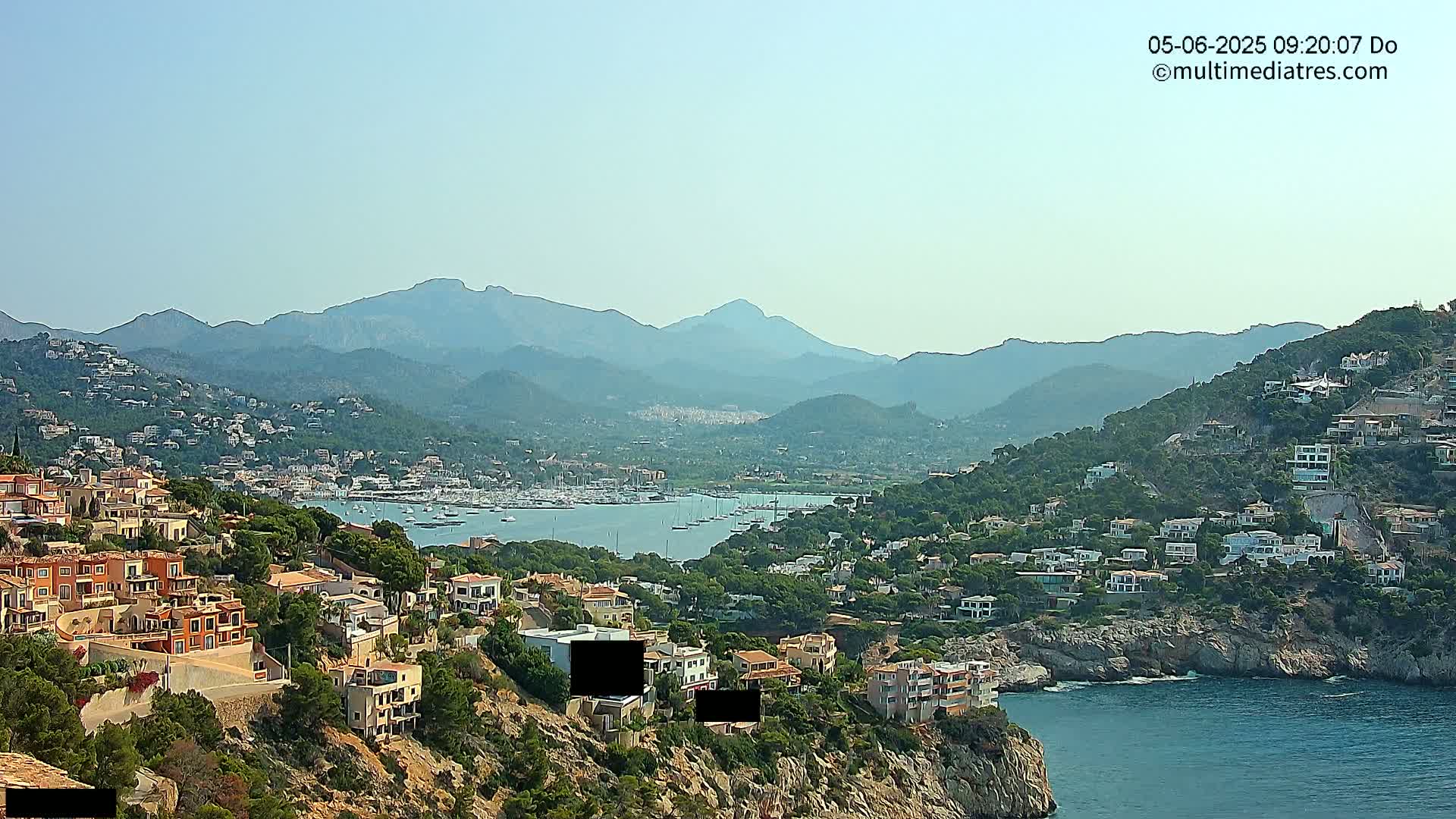 A coastal town nestled on a hillside overlooking a calm bay filled with boats, with mountains in the background under a clear sky.