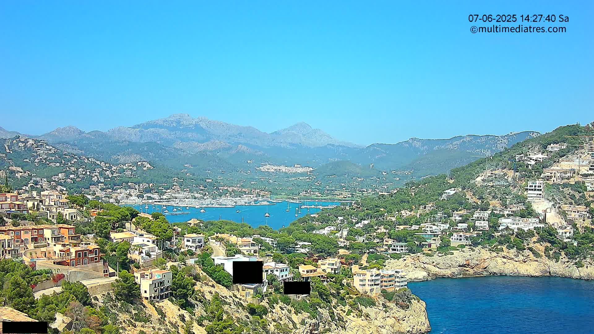A coastal town nestled against mountains overlooking a calm blue bay dotted with sailboats under a clear blue sky.