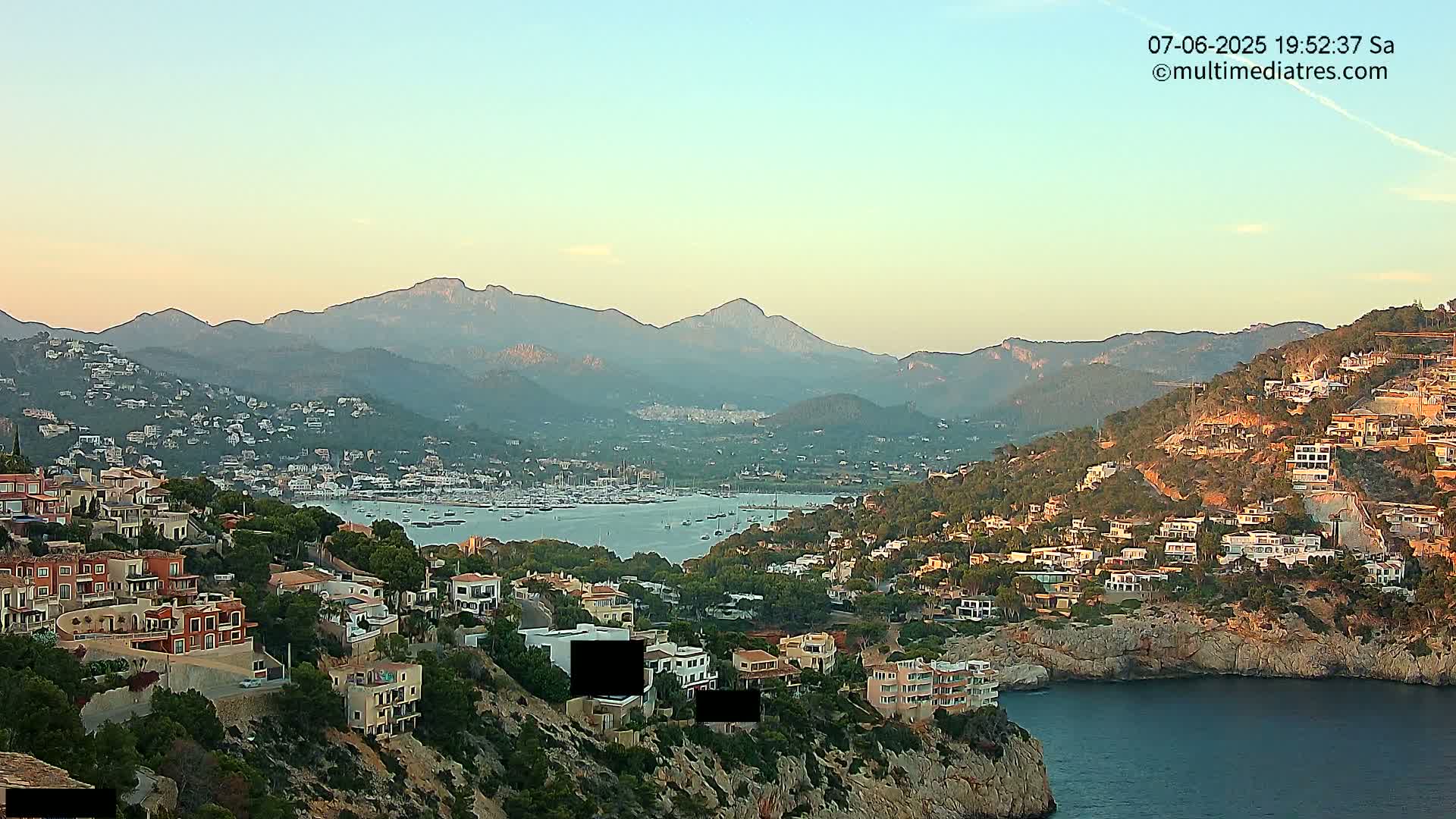 A coastal town nestled on a hillside, overlooking a calm bay filled with boats and a mountainous landscape under a clear, sunset sky.