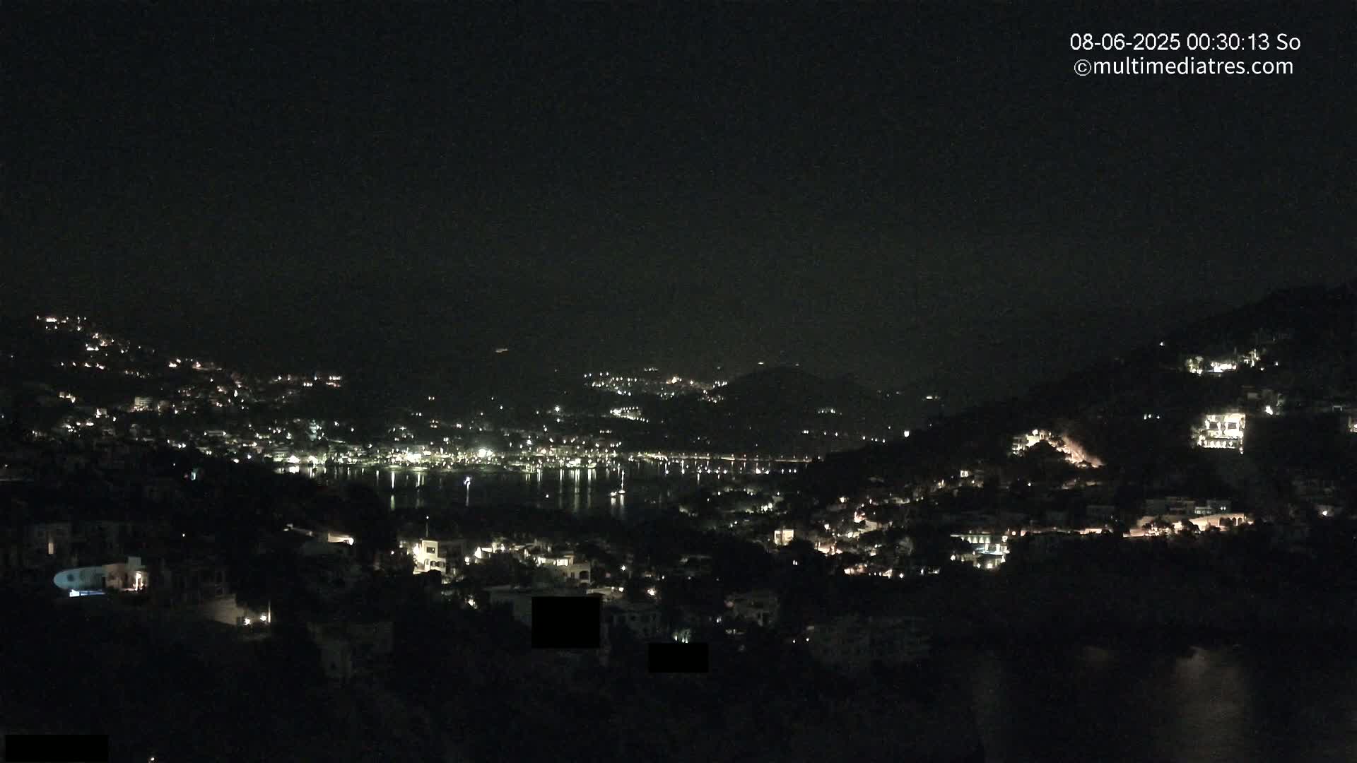 A nighttime view of a coastal town nestled on hillsides, with its lights reflecting on a calm body of water under a clear, dark sky.