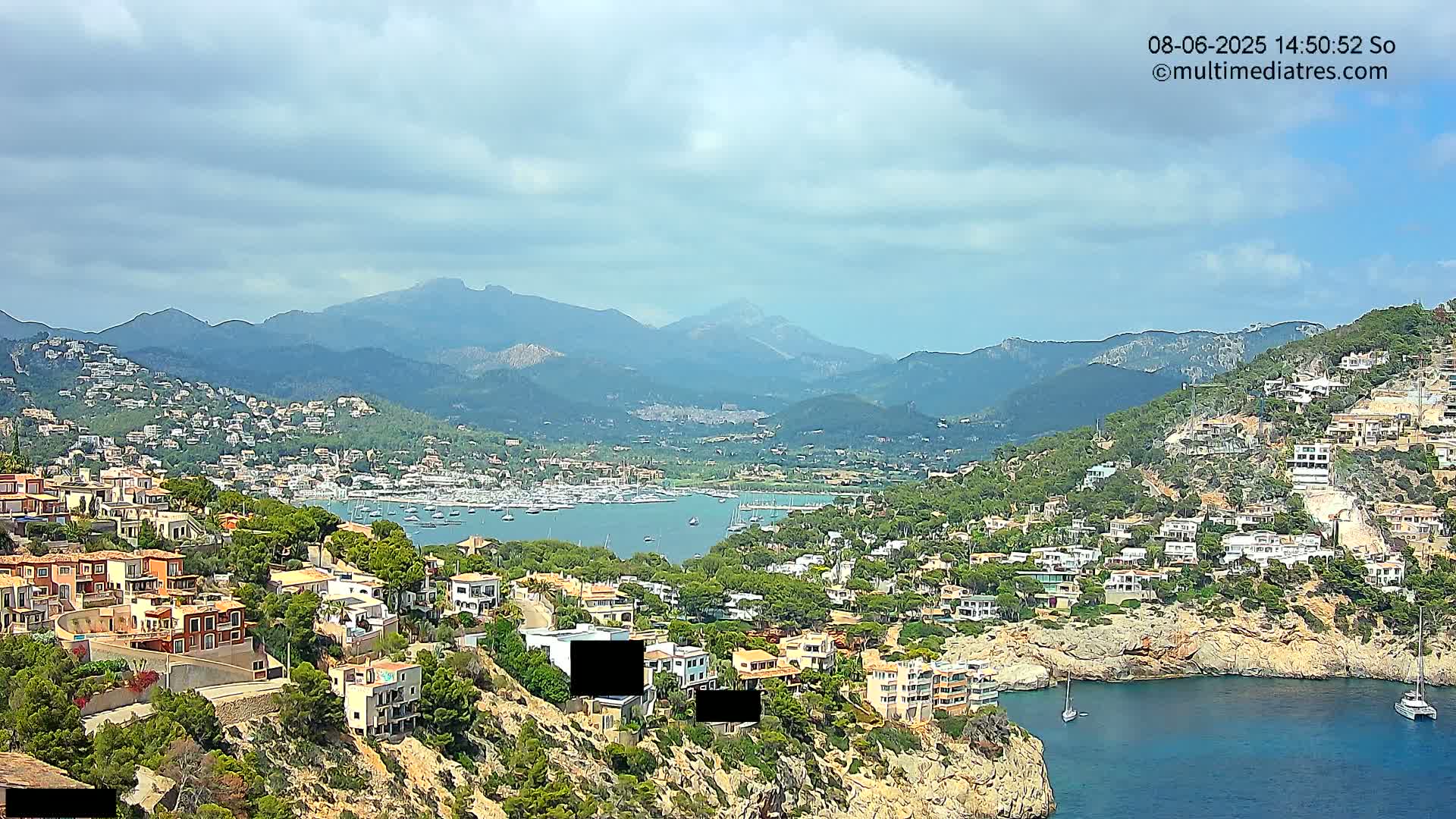 A coastal town nestled on a hillside overlooking a calm bay filled with boats, with mountains in the background under a partly cloudy sky.