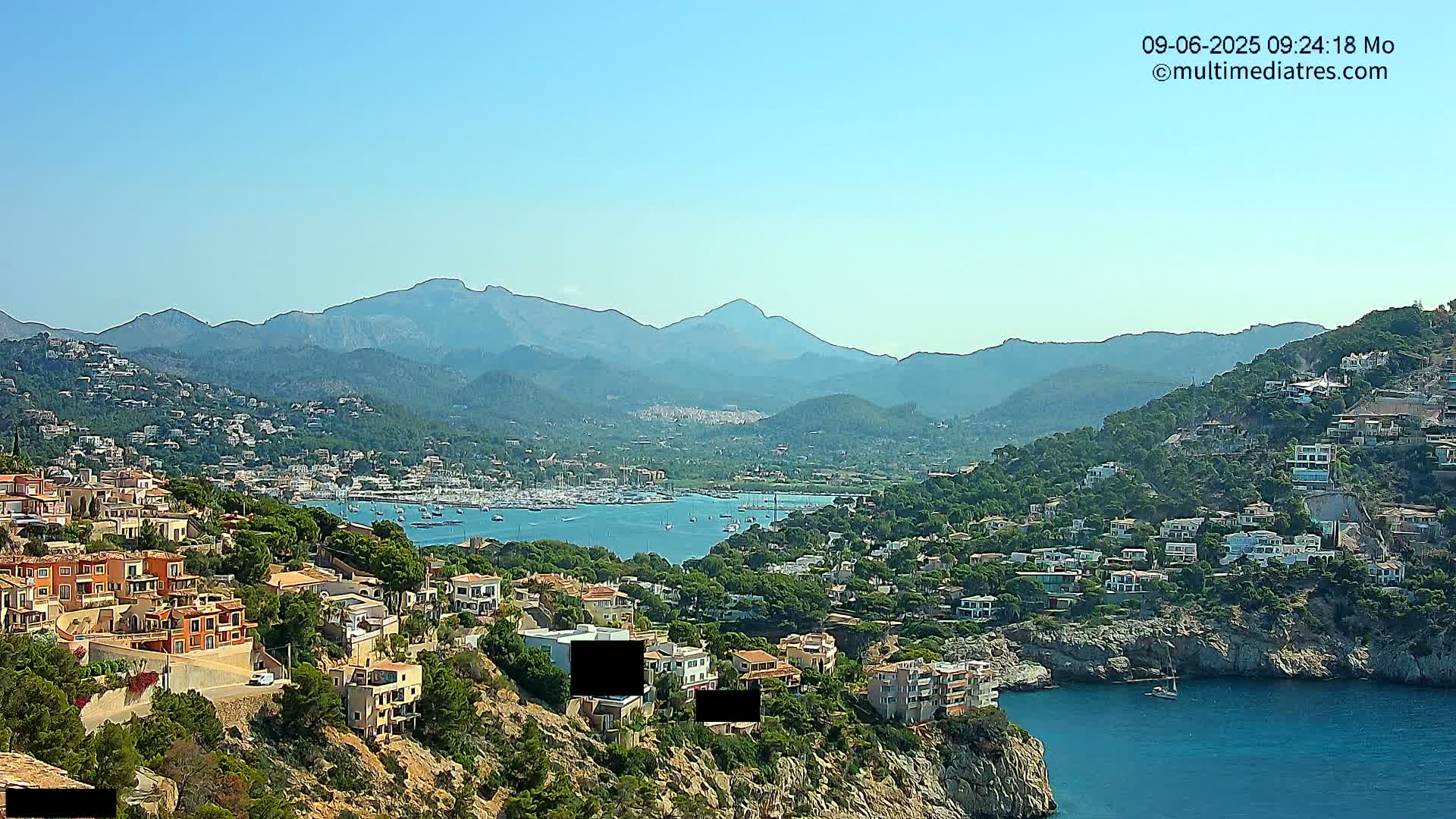 A clear, sunny day reveals a coastal town nestled on a hillside overlooking a calm bay filled with boats, all backed by a range of hazy mountains.
