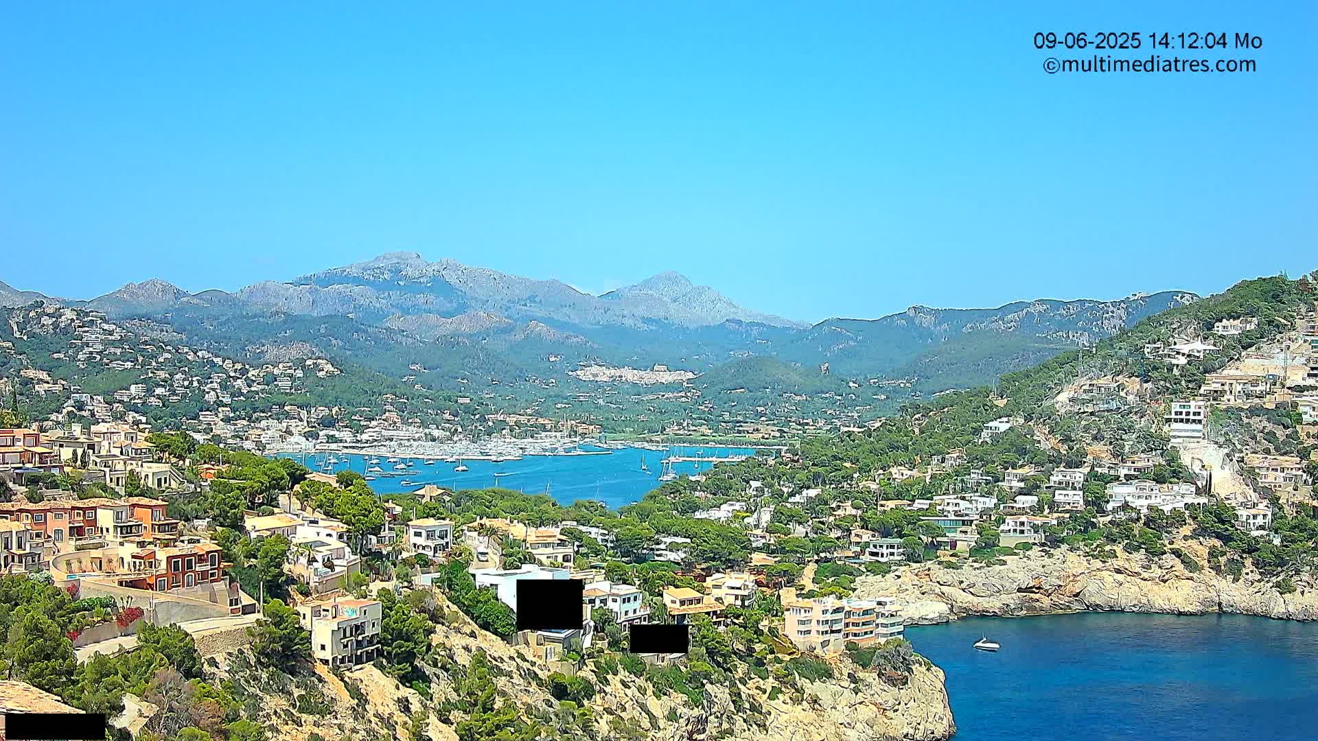 A sunny coastal town nestled in a hillside overlooking a calm bay, with mountains in the background.