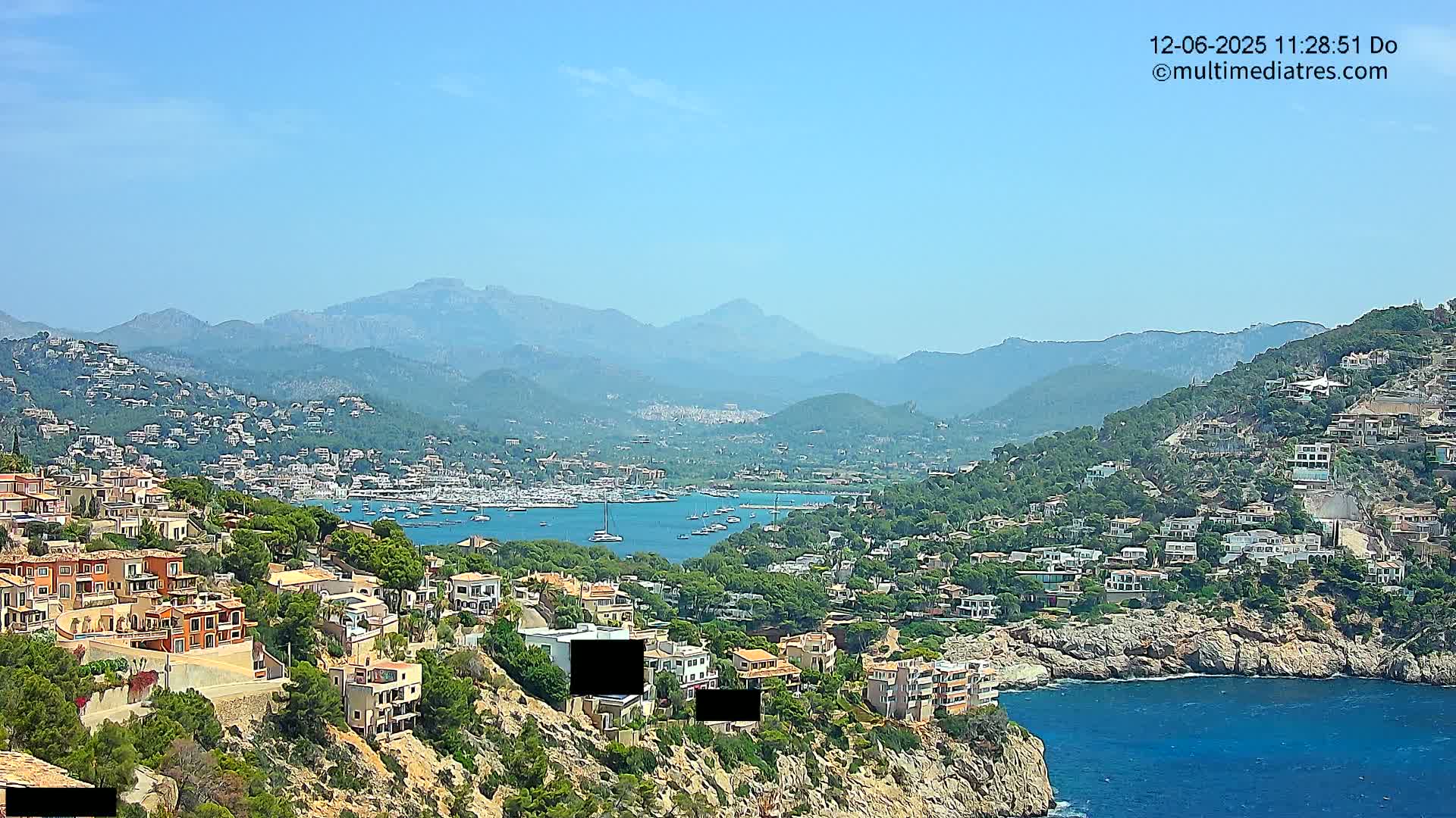 A coastal town nestled against a mountainous backdrop under a clear blue sky, with a harbor filled with boats.
