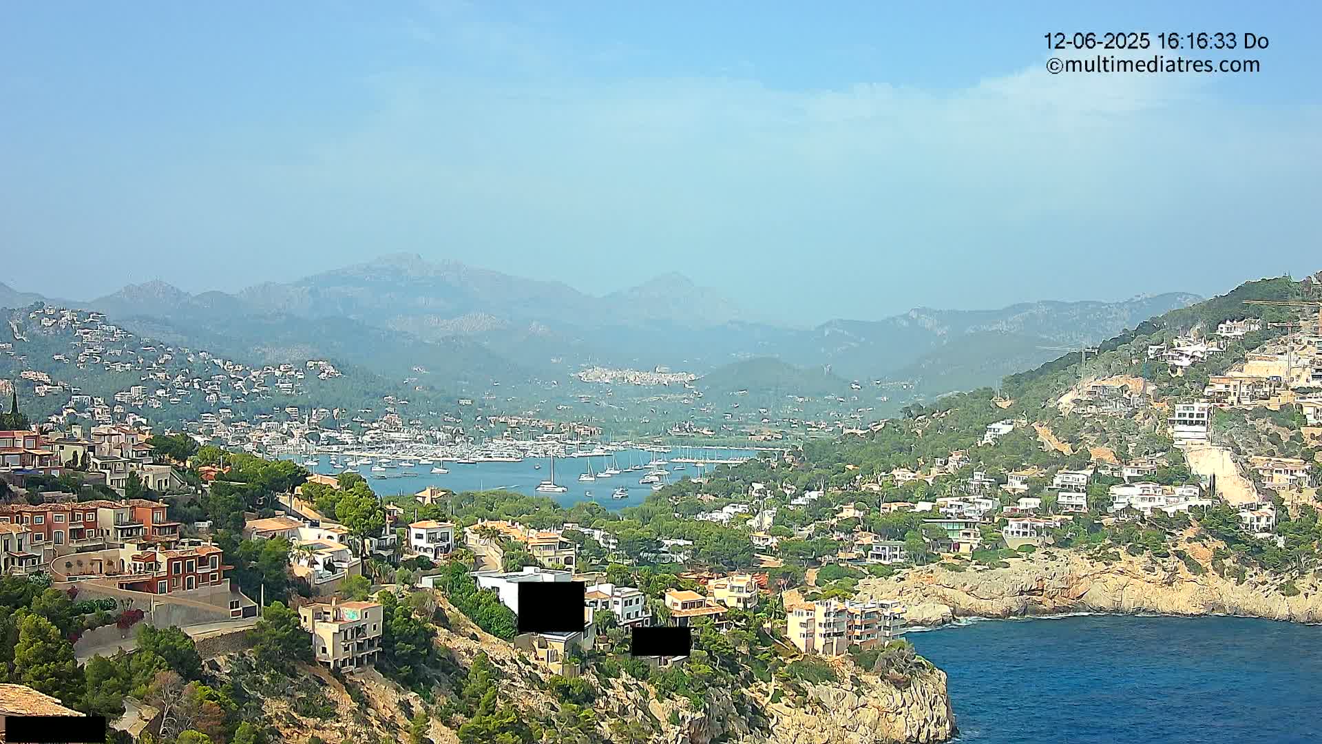 A coastal town nestled into a hillside overlooking a bay filled with boats, all under a clear, hazy sky with mountains in the background.