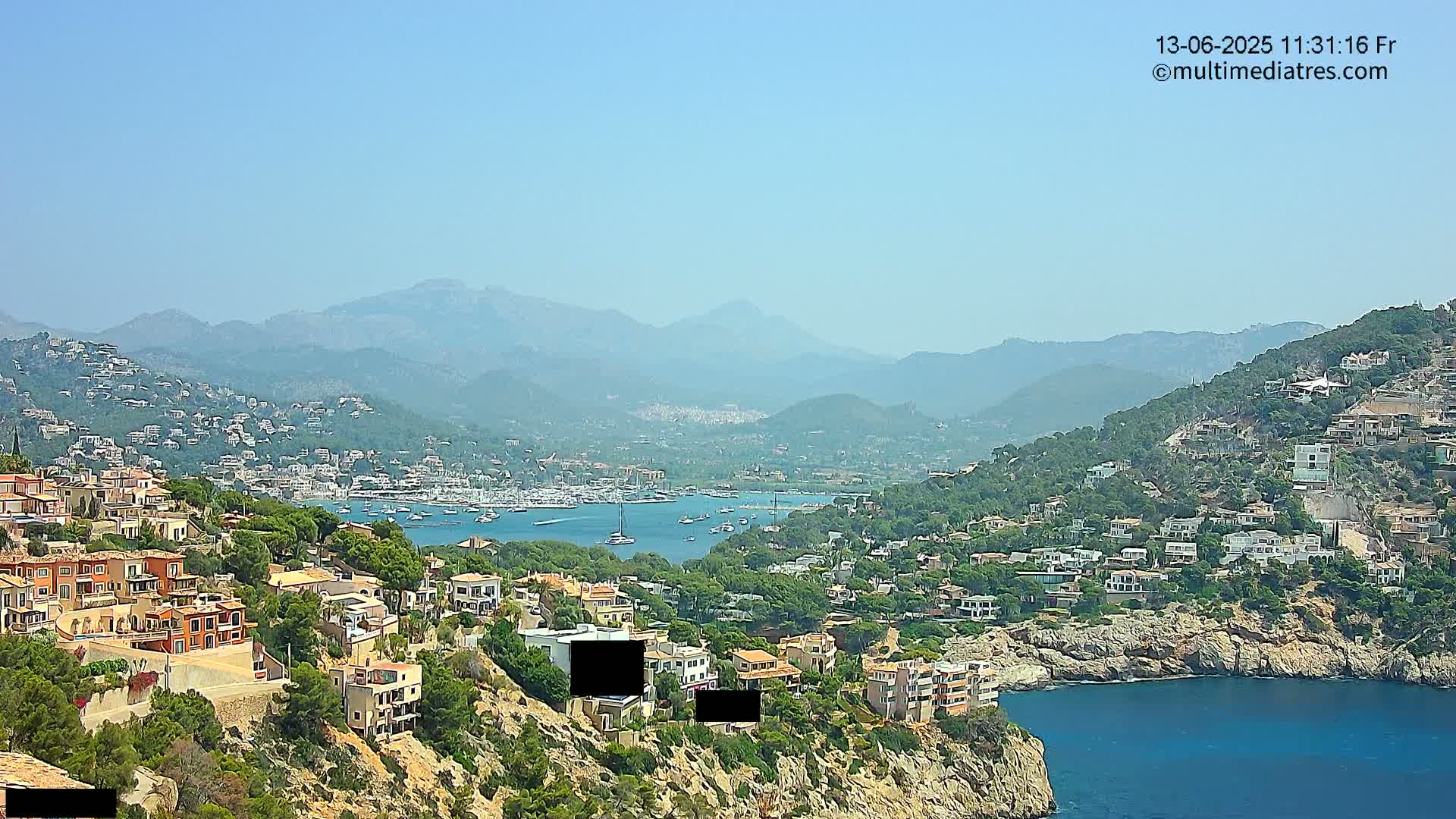 A hazy, sunny day overlooks a coastal town nestled on a hillside, with a harbor full of boats and mountains in the background.