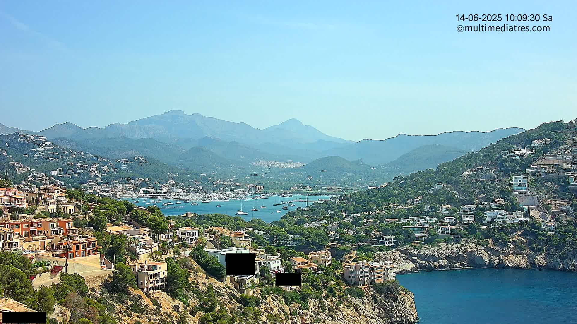 A coastal town nestled among hills and mountains under a clear blue sky overlooks a calm bay filled with boats.