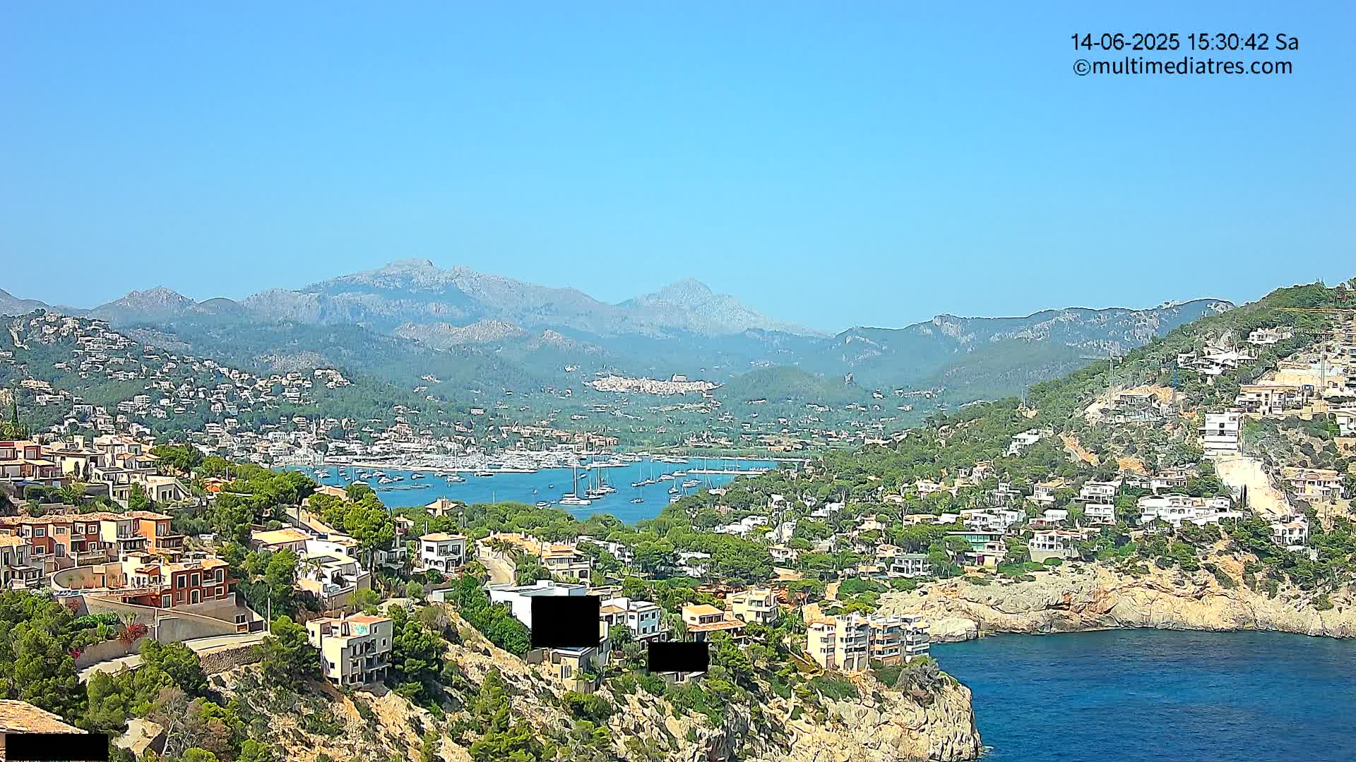 A coastal town nestled on a hillside overlooking a bay filled with boats, with mountains in the background under a clear blue sky.