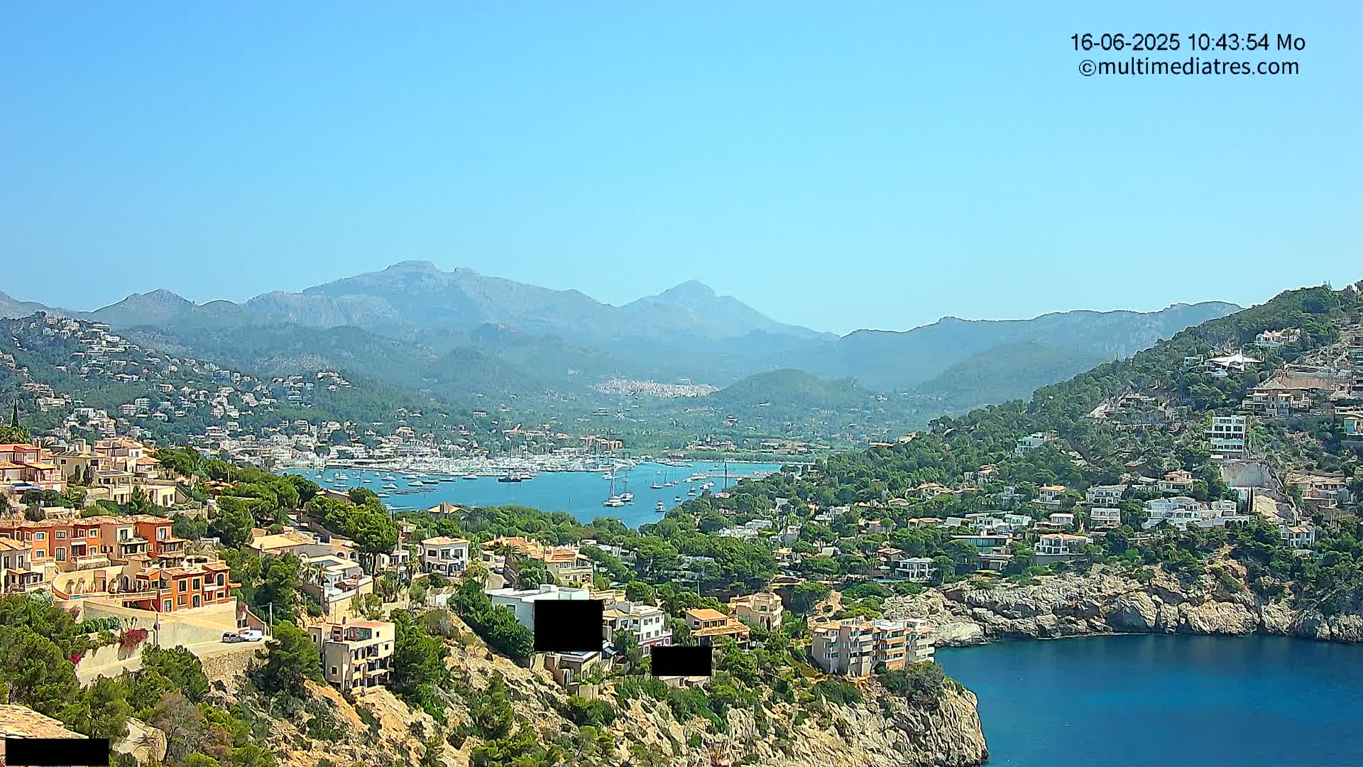 A coastal town nestled into a hillside overlooking a calm bay filled with sailboats, backed by a range of mountains under a clear blue sky.