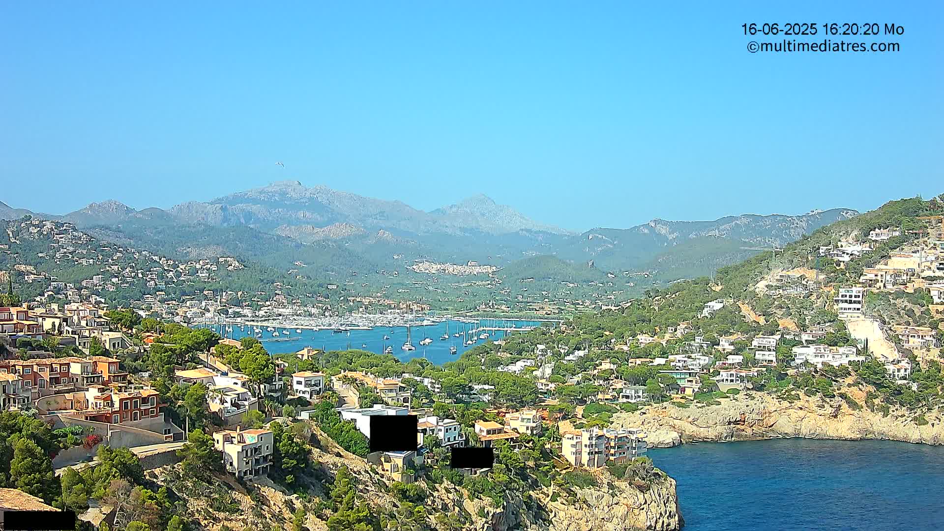 A coastal town nestled in a hillside overlooking a harbor filled with sailboats, under a clear blue sky and sunny weather, with mountains in the background.
