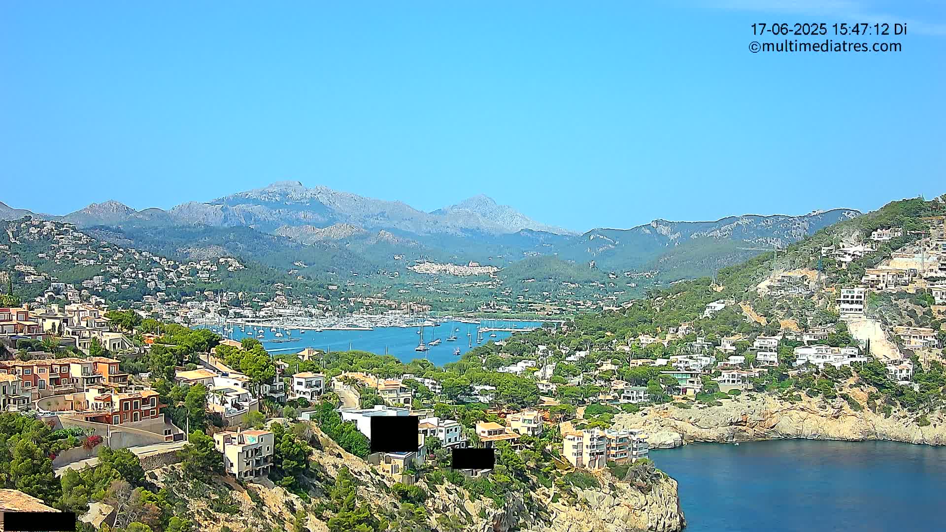 A clear, sunny day reveals a coastal town nestled on a hillside overlooking a calm bay filled with sailboats, all backed by a range of mountains.