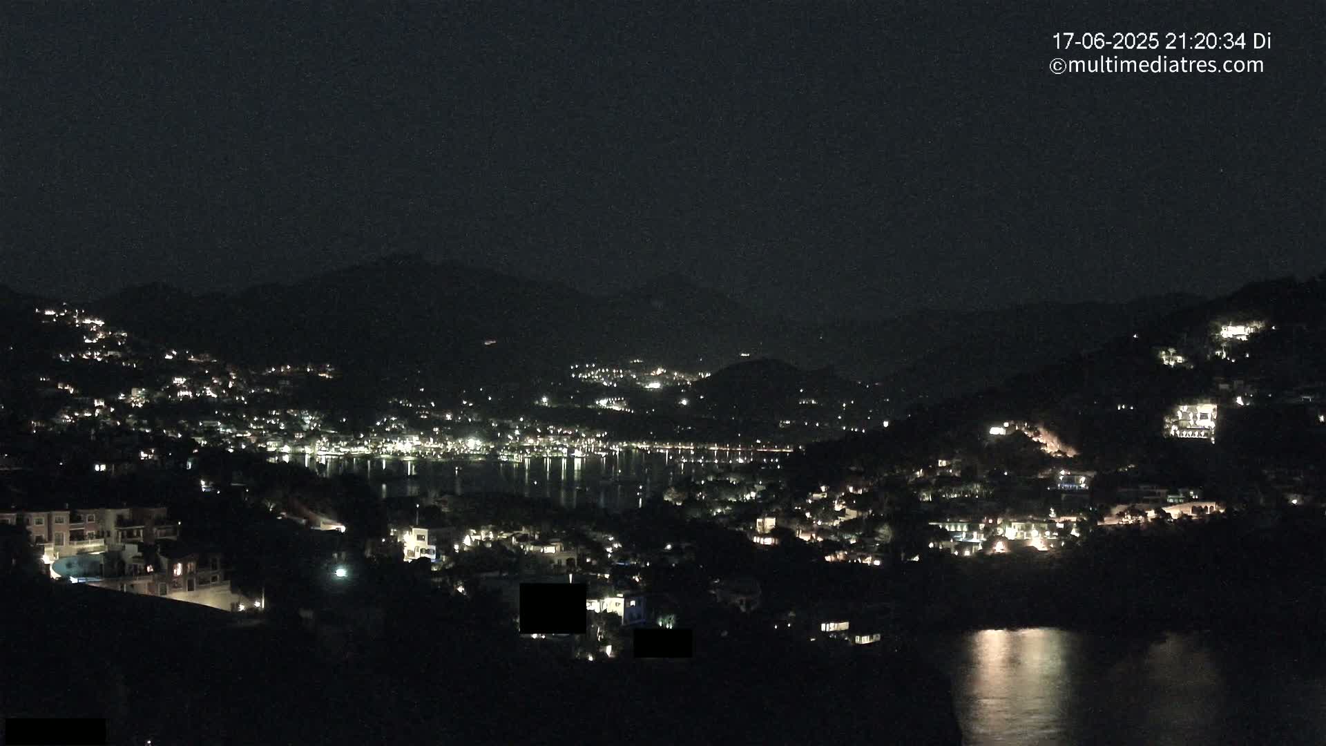 A nighttime view of a coastal town nestled between dark hills, with the lights of buildings and a calm body of water reflecting the lights.