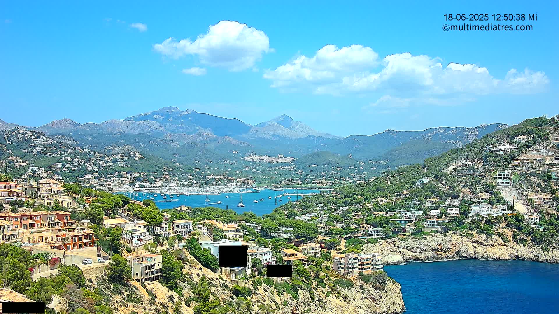 A coastal town nestled into a hillside, with a harbor full of boats and mountains in the background under a partly cloudy sky.