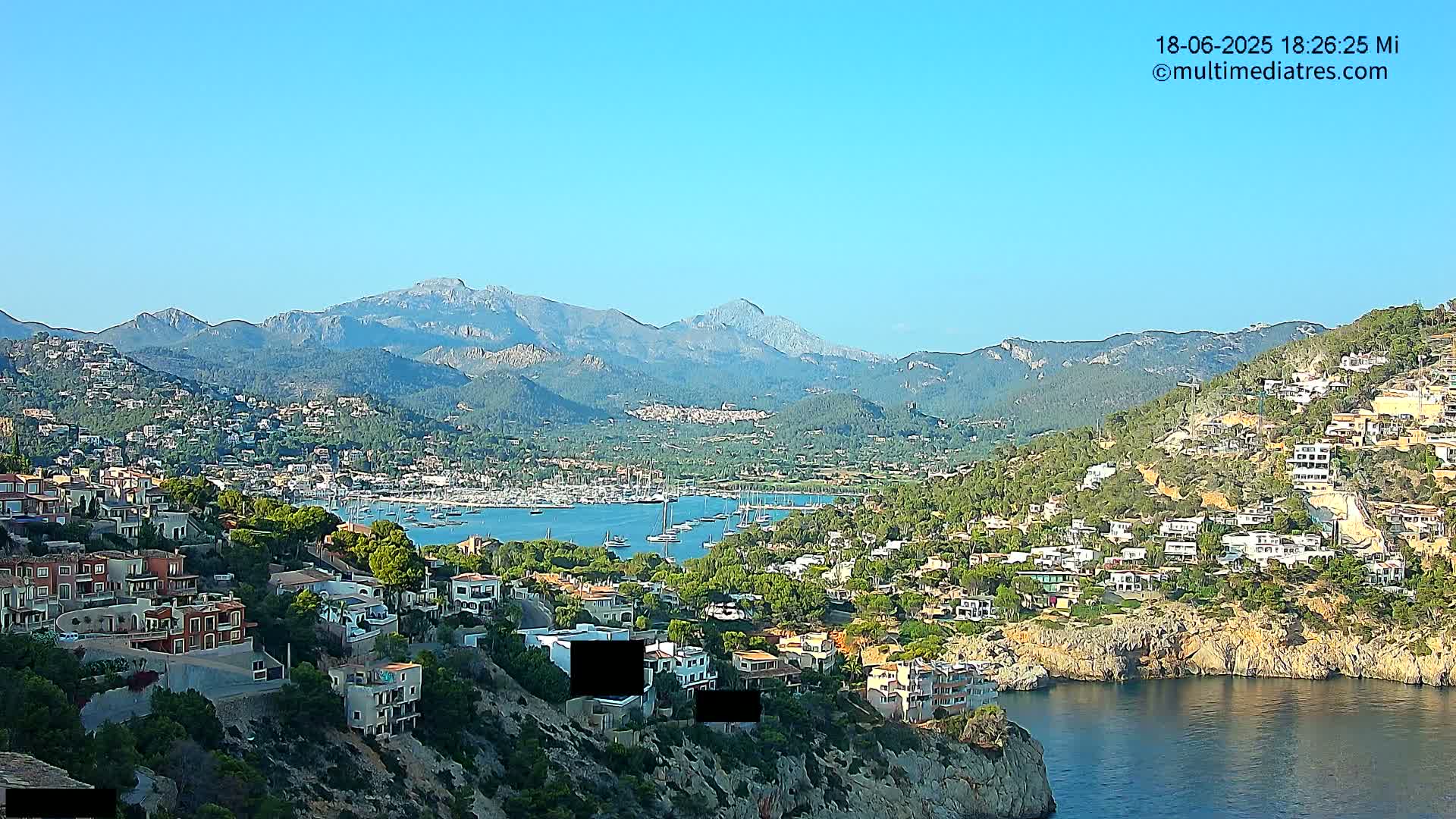 A coastal town nestled against a mountain range under a clear blue sky overlooks a harbor filled with sailboats.