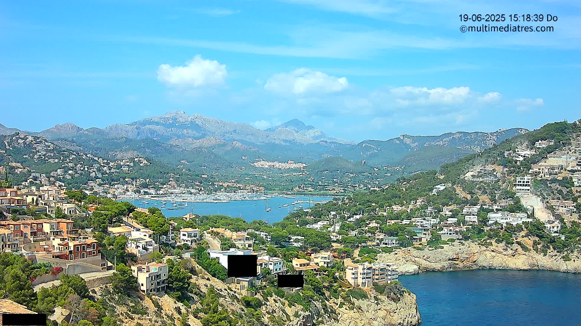 A coastal town nestled in a mountainous region with a clear blue sky and scattered clouds, featuring a harbor filled with boats.