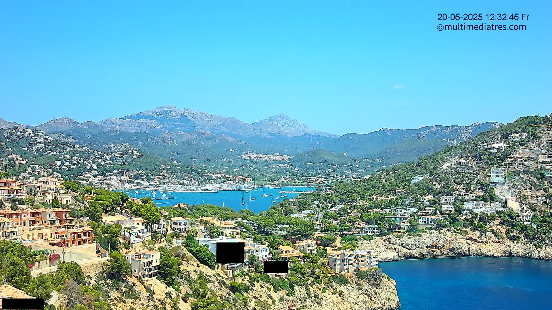 A sunny coastal town nestled on a hillside overlooking a calm bay filled with sailboats, with mountains in the background.