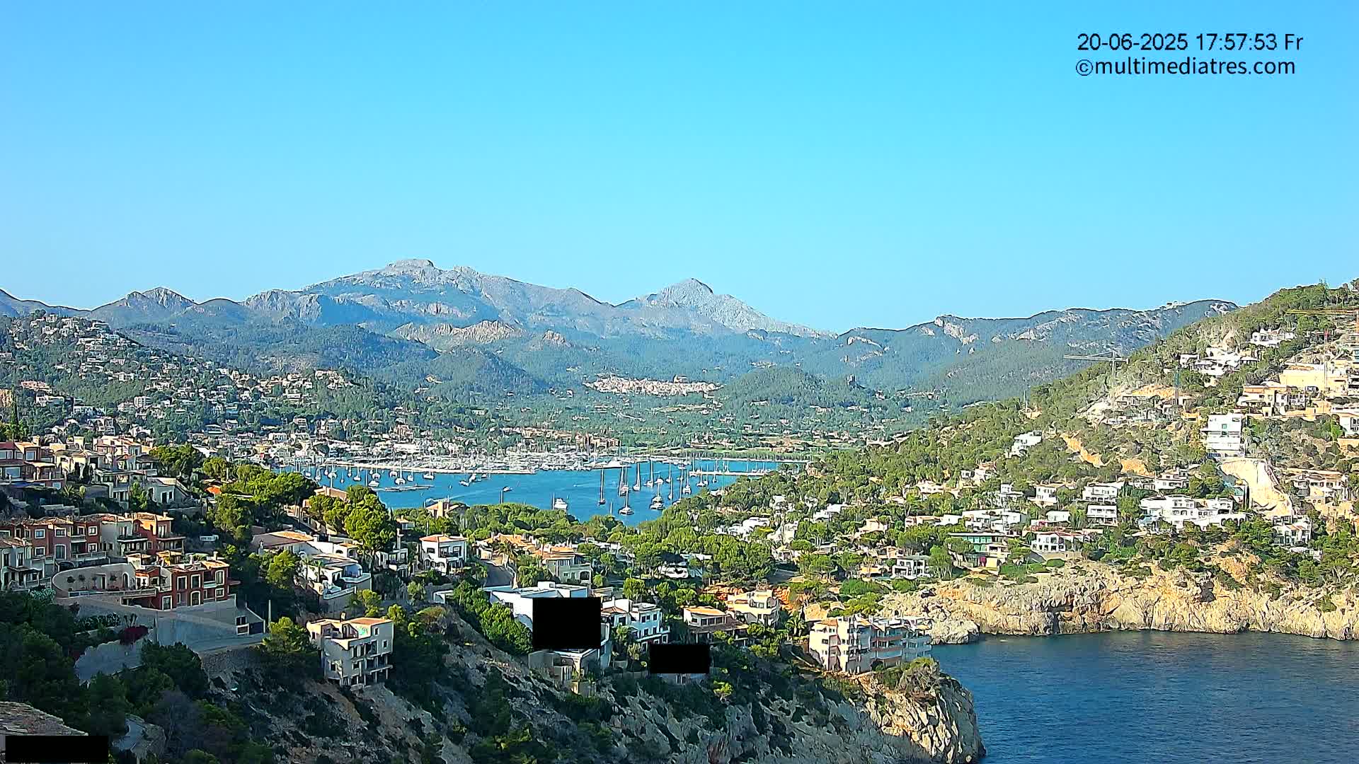 A coastal town nestled against mountains under a clear blue sky, with a harbor filled with sailboats.